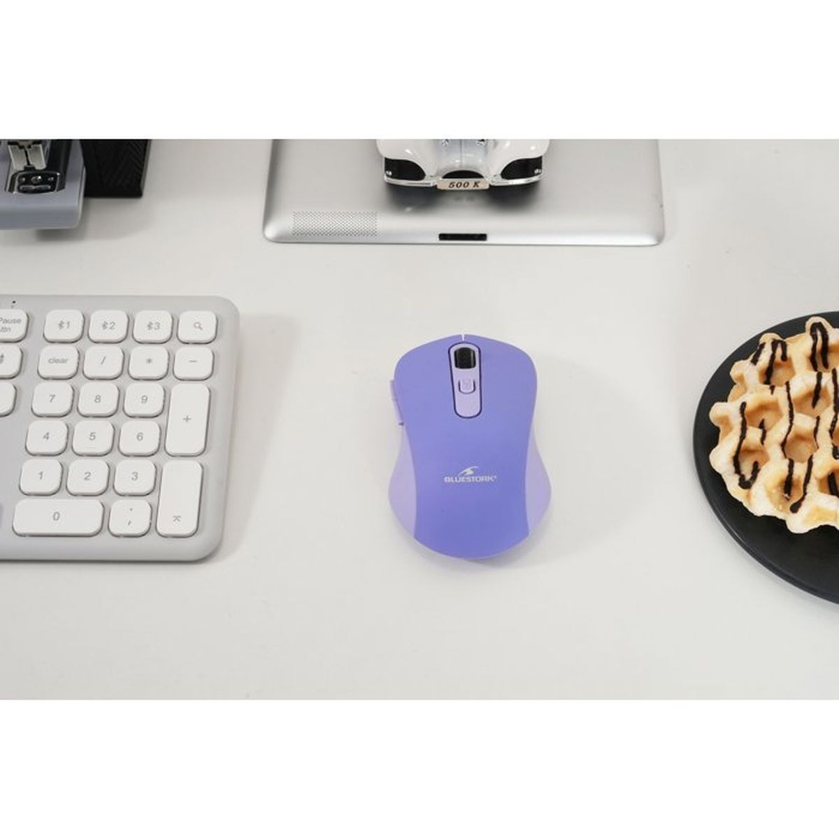 Un ratón de ordenador morado en un escritorio blanco con un teclado, una tableta y un gofre en un plato.