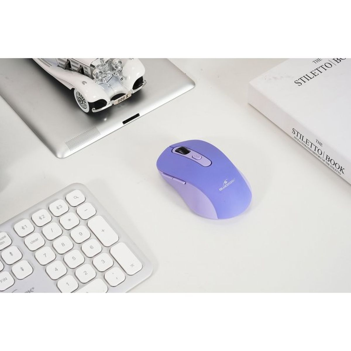 Un ratón de ordenador morado sobre un escritorio blanco junto a un teclado, una tableta con un coche de juguete y un libro.