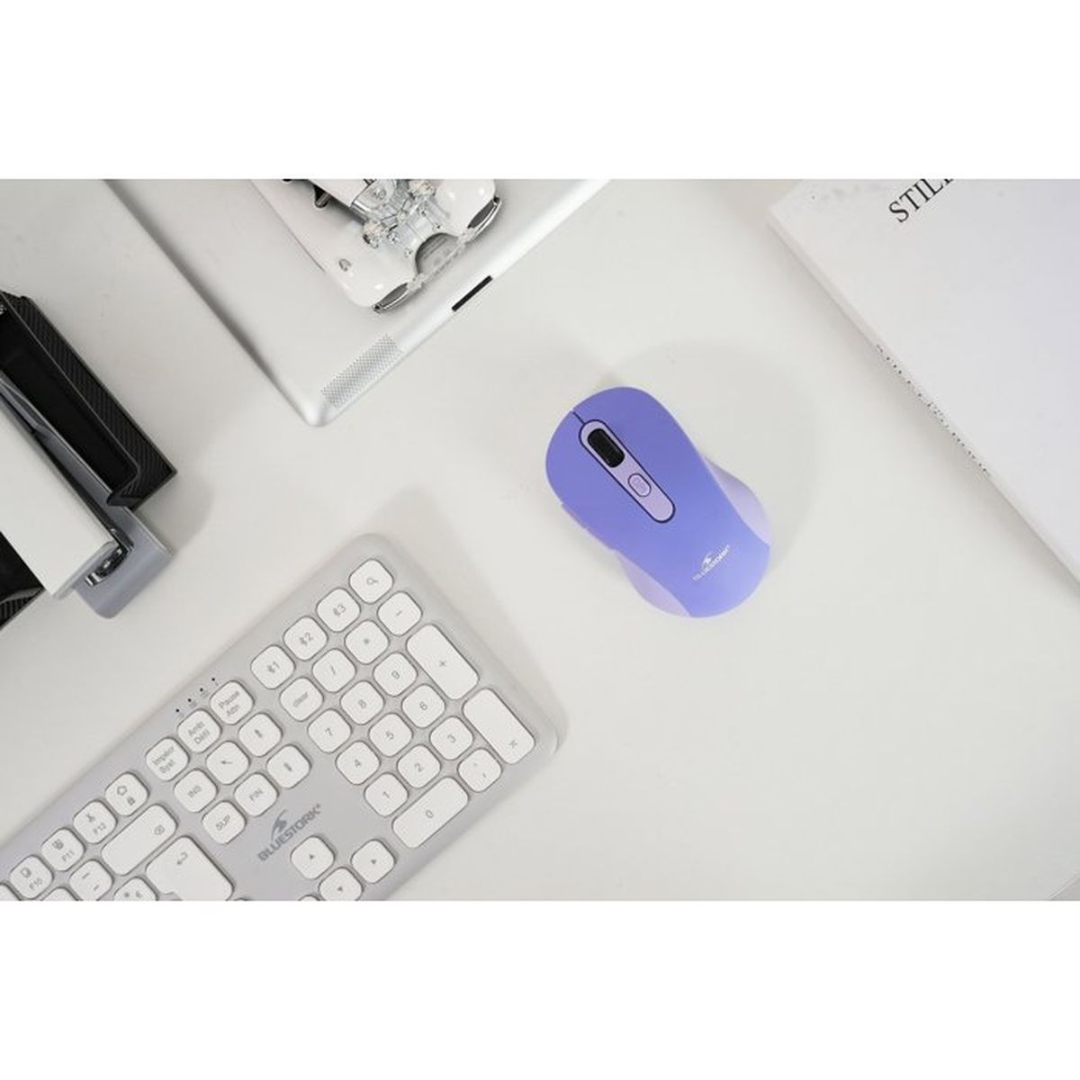 Un ratón de ordenador morado descansa sobre un escritorio blanco junto a un teclado, tableta y libro.