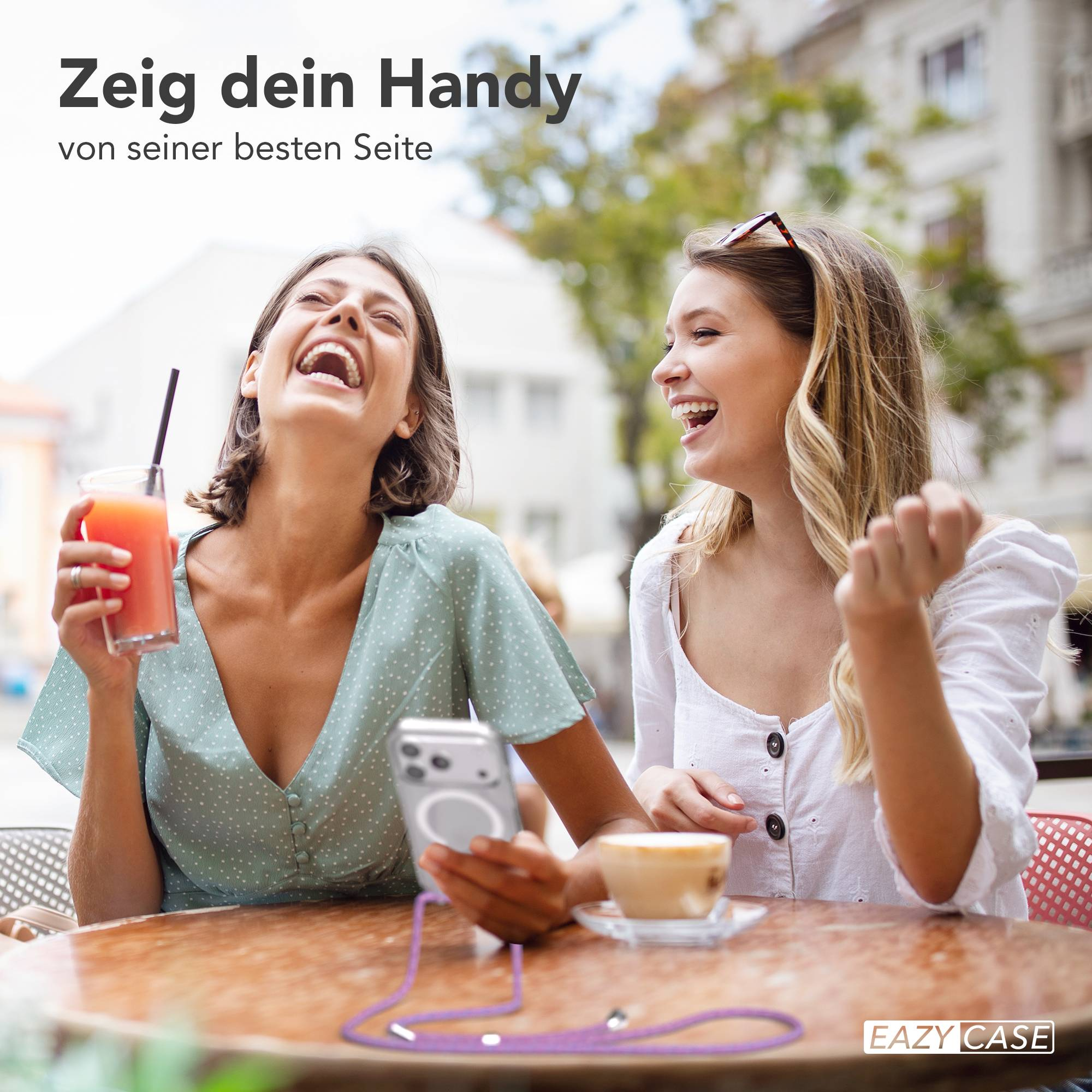 Zwei junge Frauen lachen gemeinsam an einem Outdoortisch, halten Getränke und ein Smartphone.