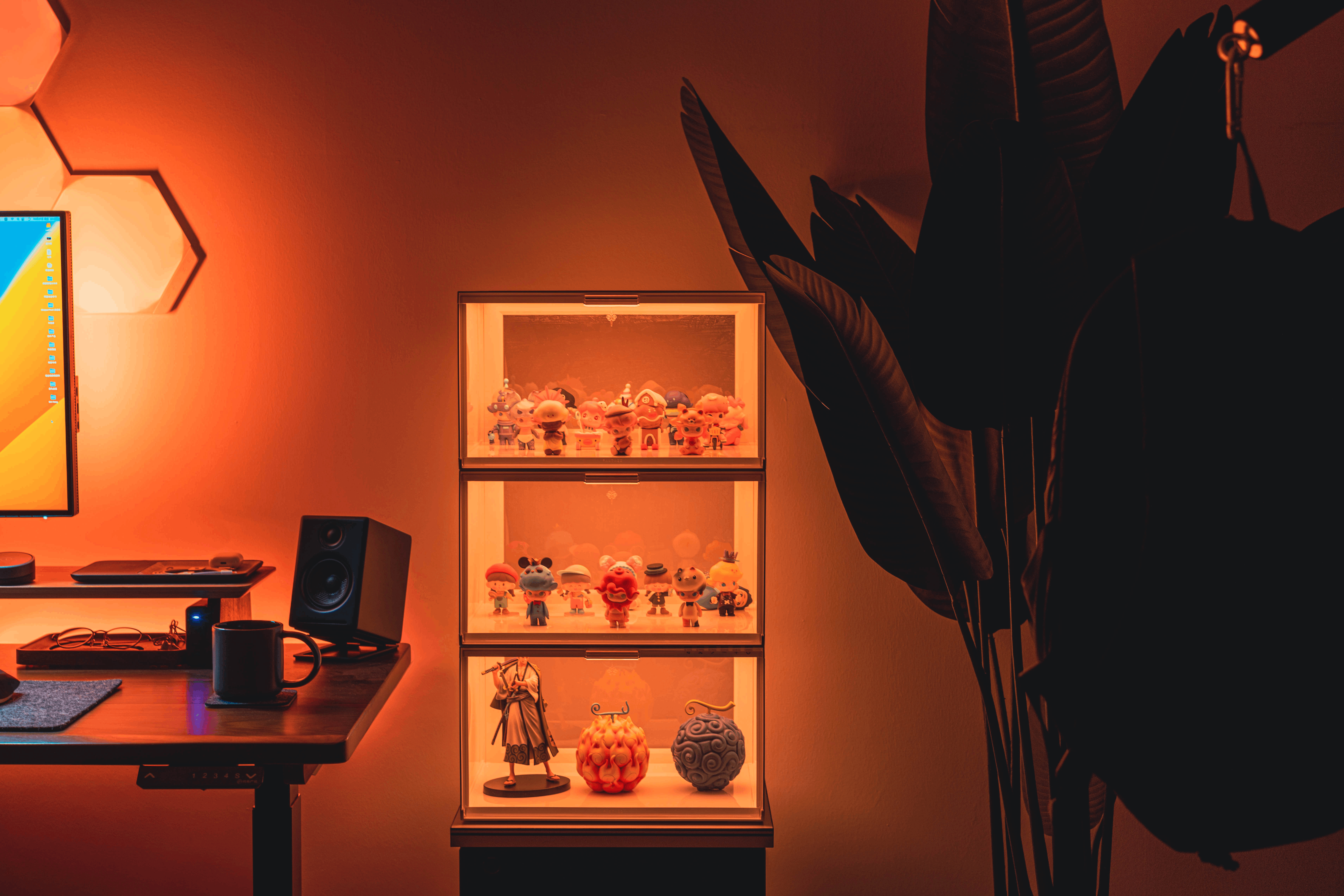 Eine beleuchtete Vitrine mit kleinen Spielfiguren steht neben einem Schreibtisch und einer großen Pflanze.