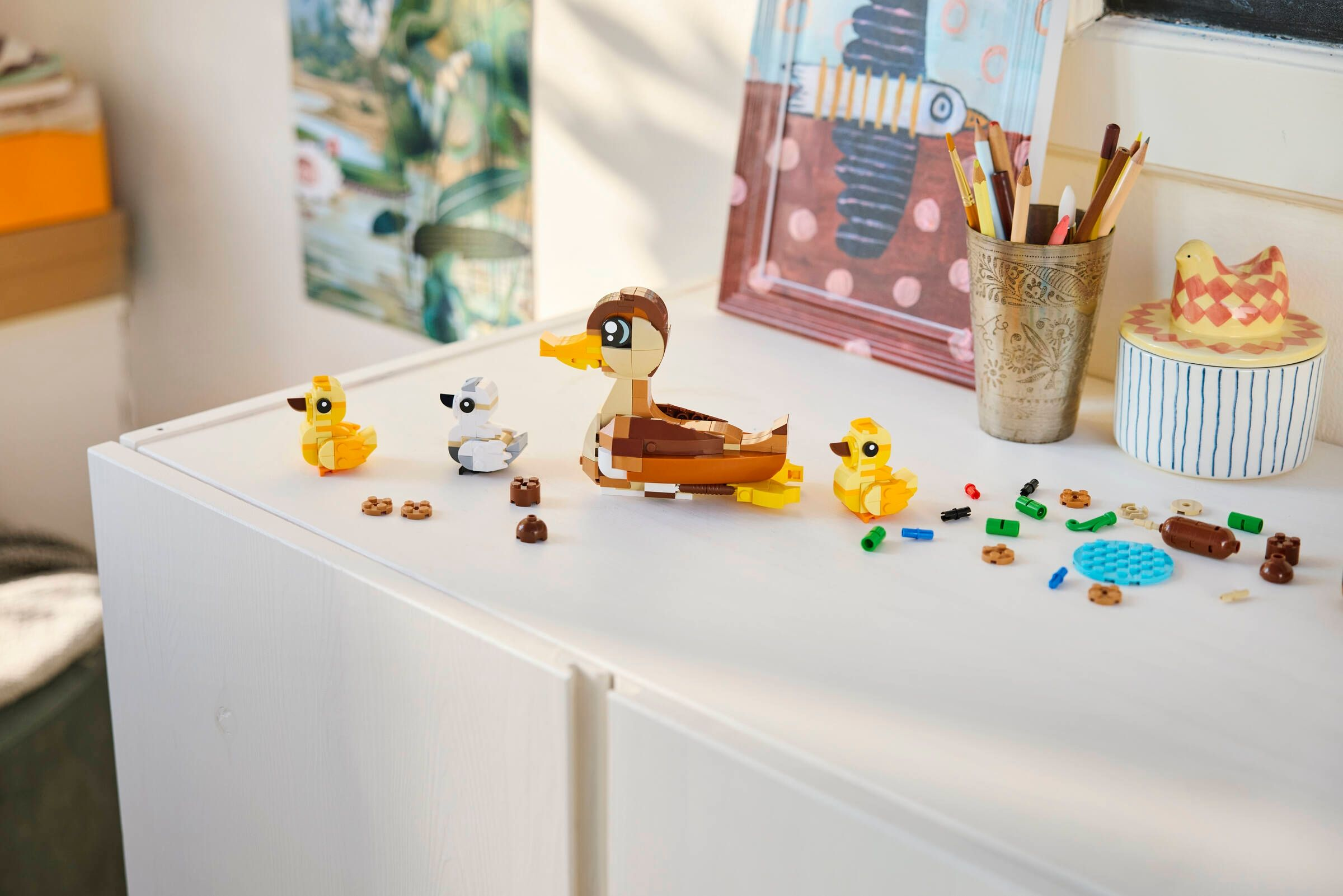 Des modèles de canards Lego de différentes couleurs et tailles se trouvent sur une commode blanche à côté de crayons.