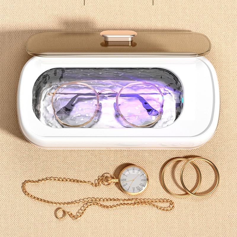 Eine UV-Desinfektionsbox enthält eine Brille. Davor liegen eine Taschenuhr mit Kette und zwei Ringe auf beigem Stoff.