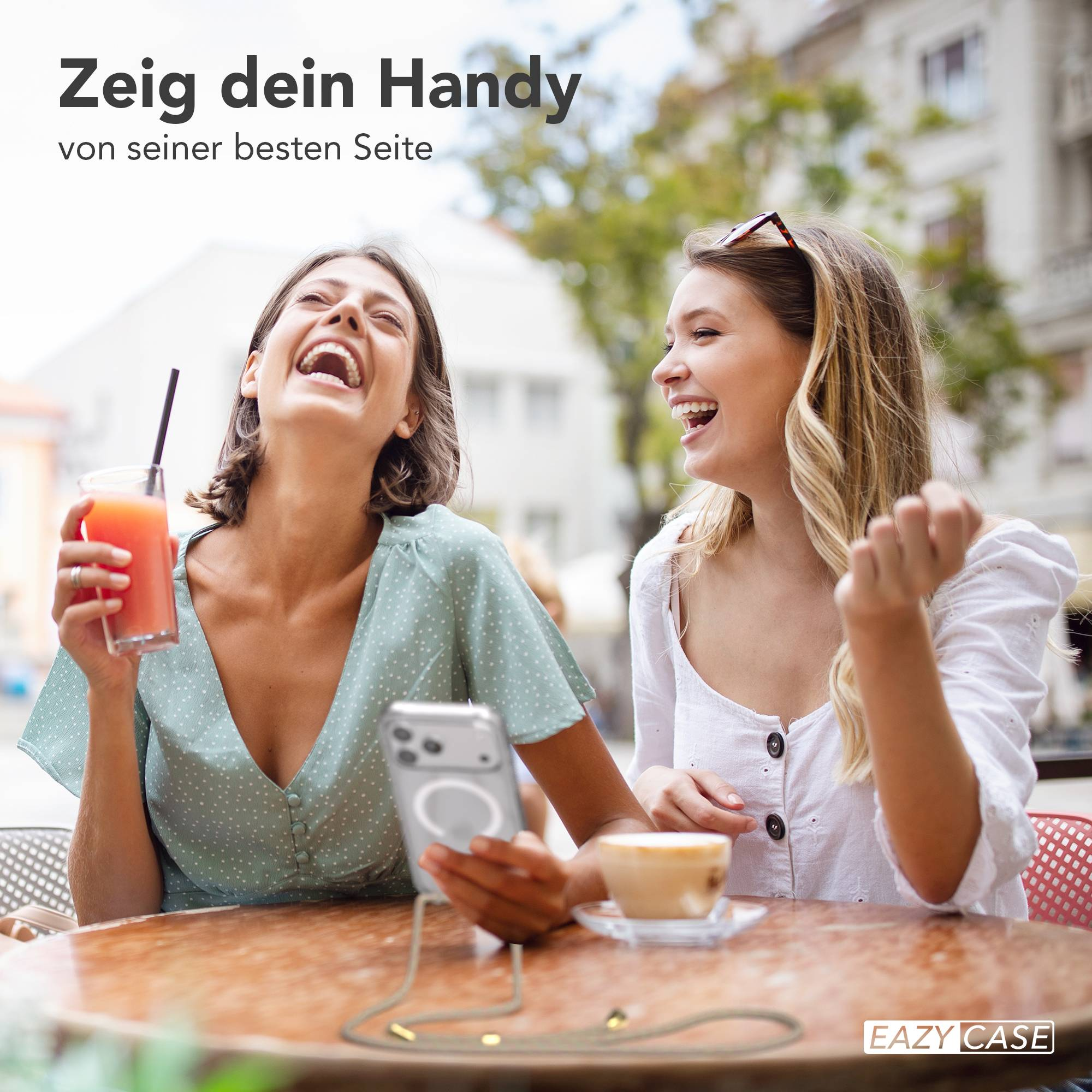 Zwei junge Frauen lachen an einem Straßencafé-Tisch, halten ein Getränk und ein Smartphone mit Umhängeband.