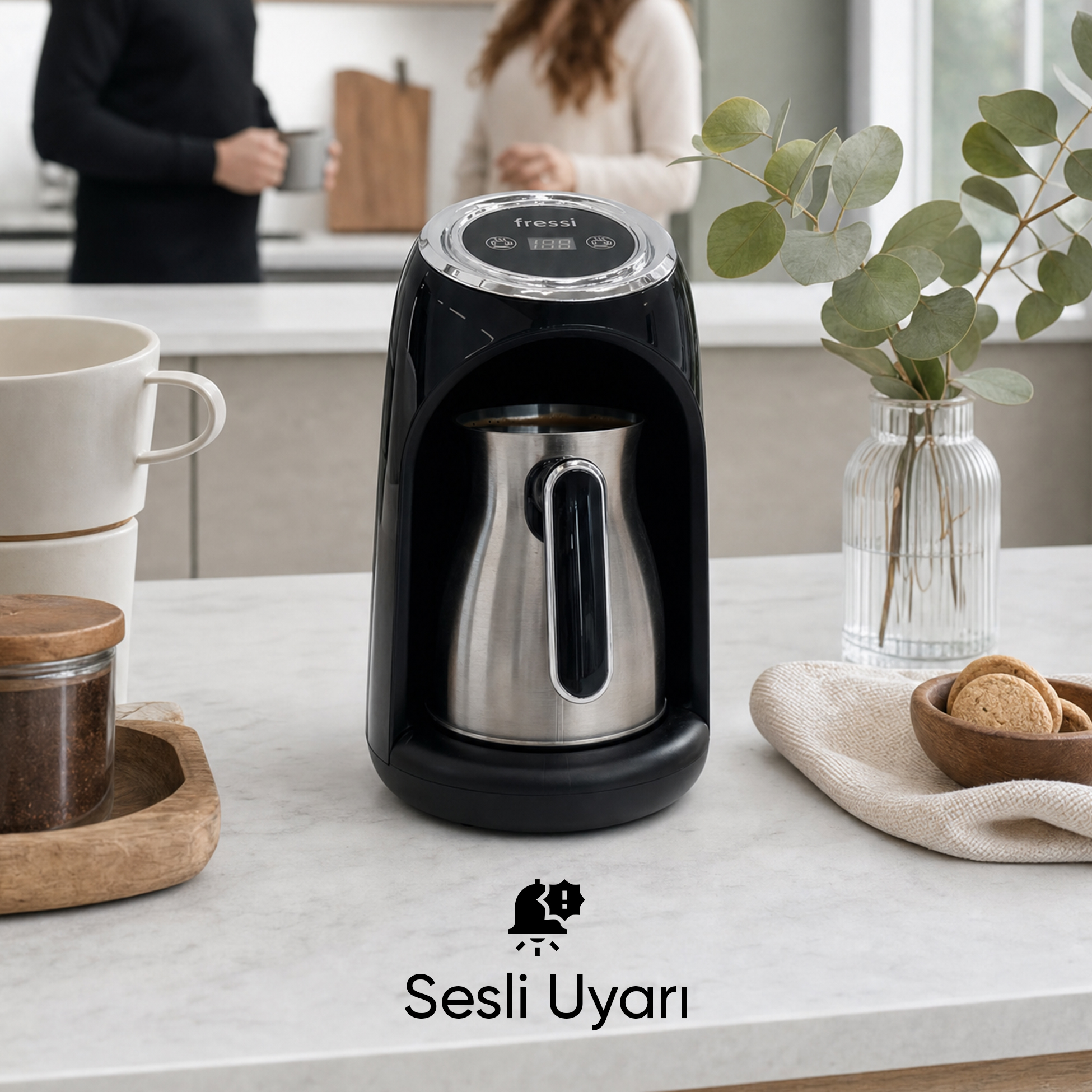 Gümüş renkli cezveli siyah bir kahve makinesi, kurabiyeler ve bir bitkinin yanında tezgahta duruyor.