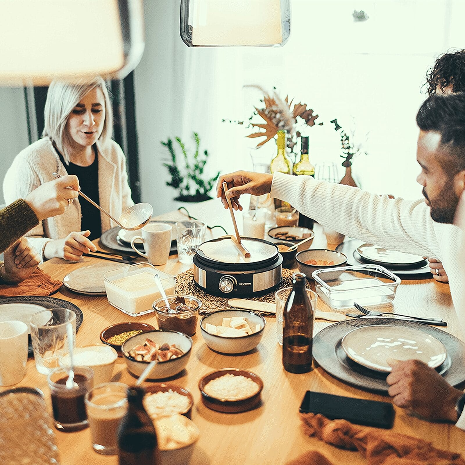 Eine diverse Gruppe von Freunden sitzt an einem Holztisch und genießt ein Raclette-Essen mit Beilagen.