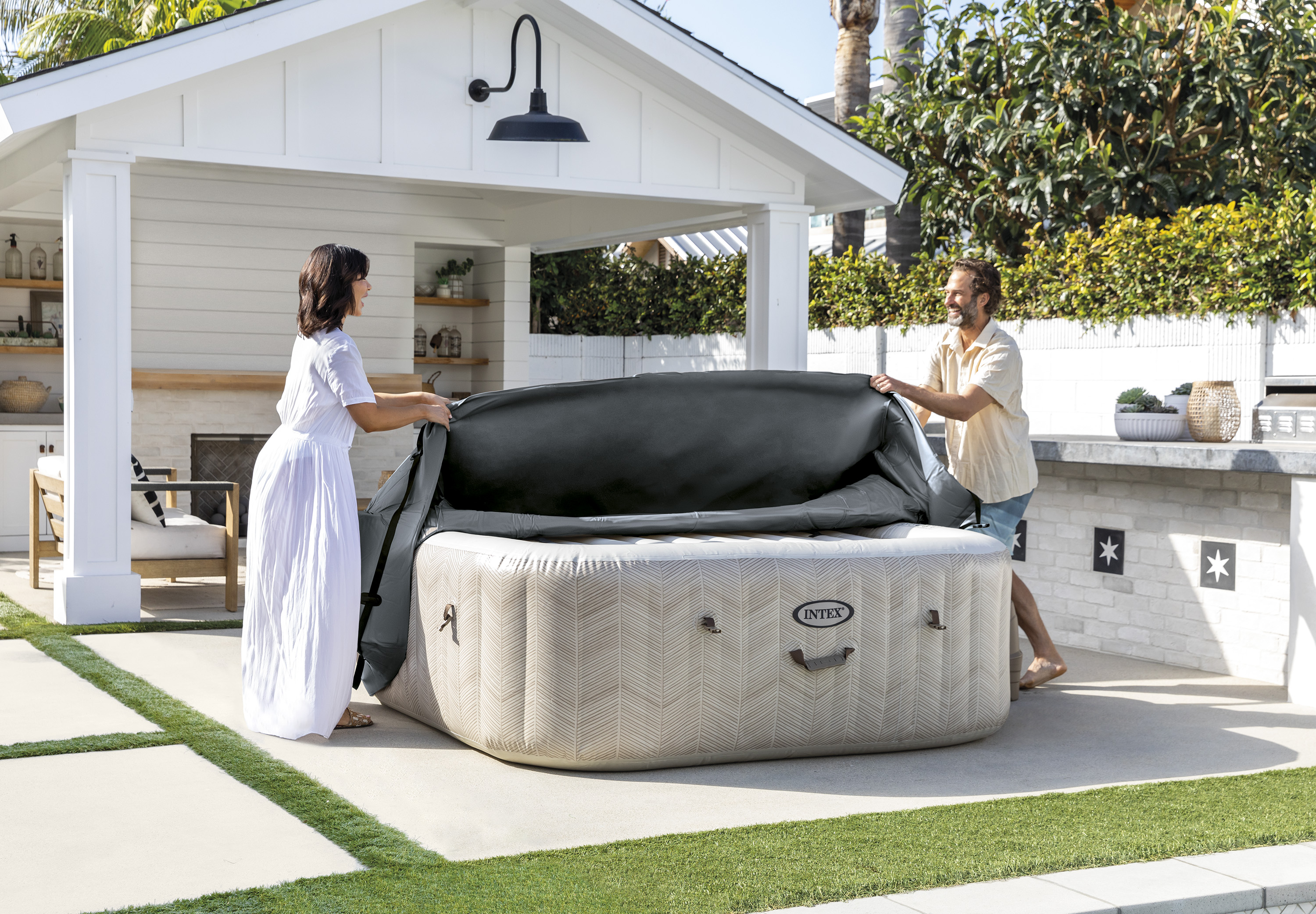 Ein Paar öffnet die graue Abdeckung eines aufblasbaren Intex-Whirlpools im Garten vor einem hellen Haus.