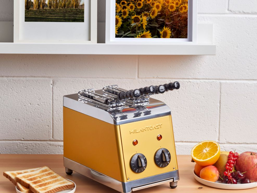 Ein goldener Retro-Toaster steht auf einem Holztisch, umgeben von Toast und frischem Obst, vor einer weißen Ziegelwand.