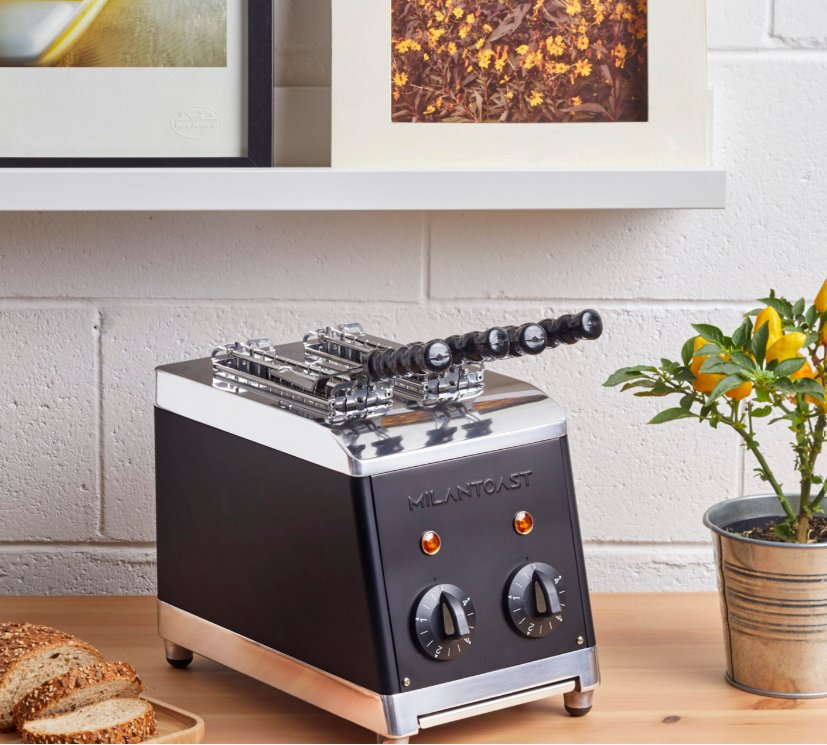Ein schwarzer Milantoast-Toaster mit Chromdetails steht auf einem Holztisch vor einer weißen Backsteinwand.
