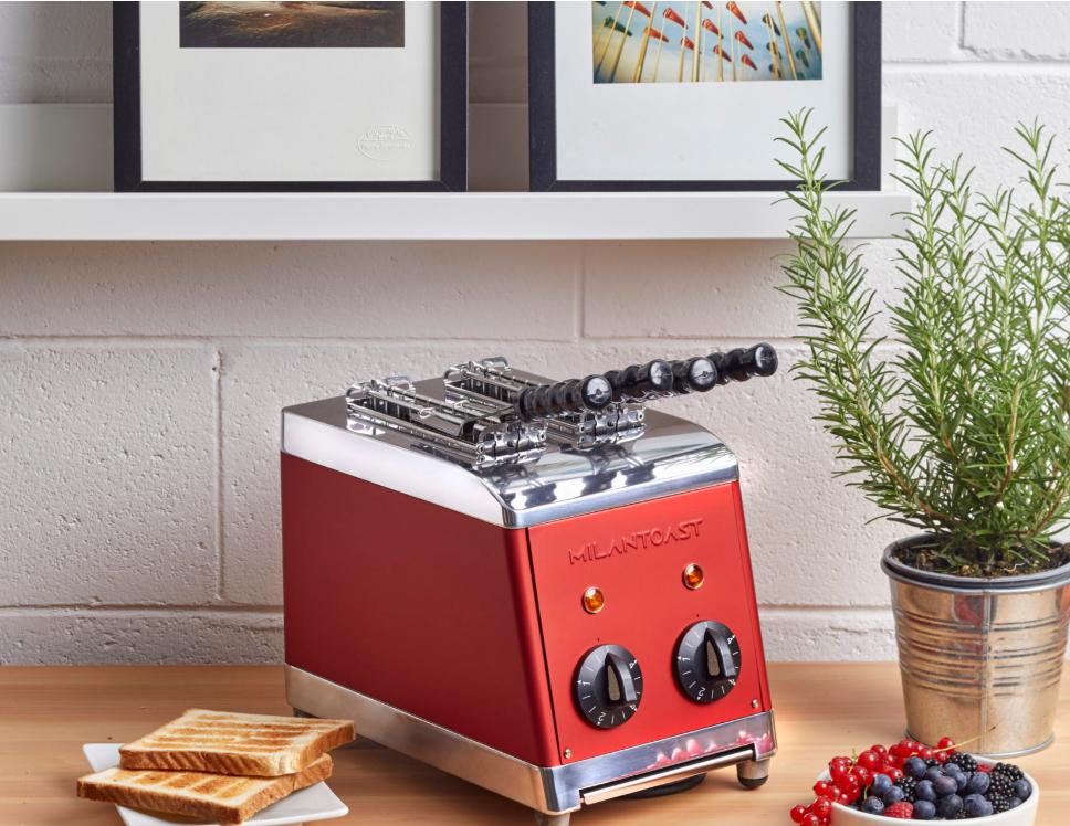 Ein roter und chromfarbener Retro-Milantoast-Toaster auf einem Holztisch mit Toast, Beeren und einer Topfpflanze.