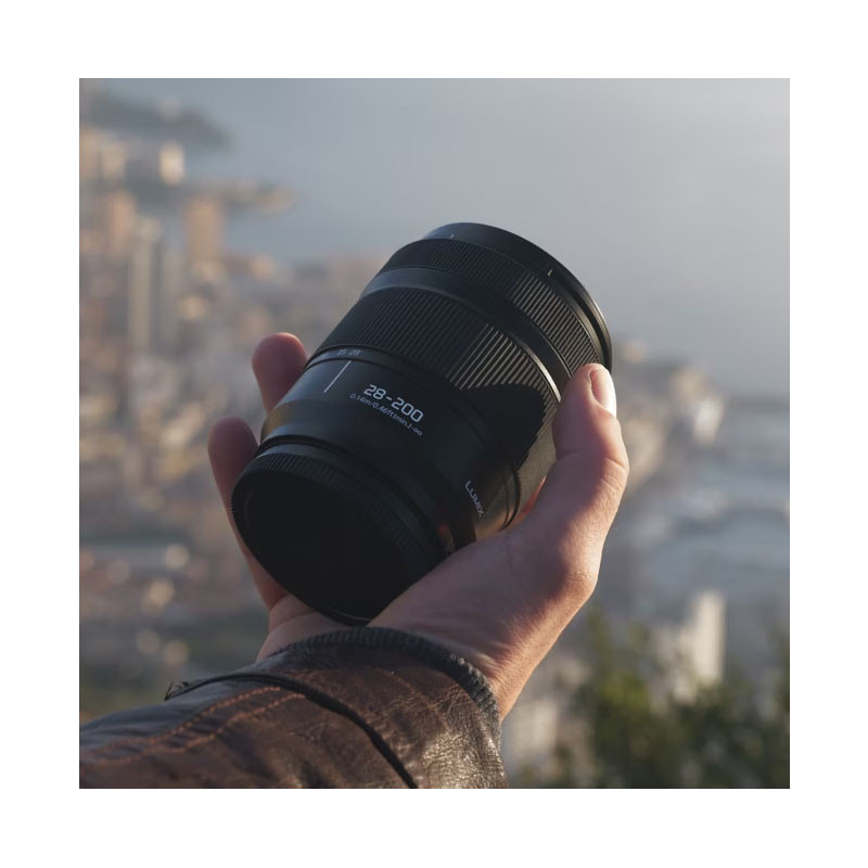 Una persona tiene in mano un obiettivo fotografico nero con etichetta 28-200 e sfondo sfocato.