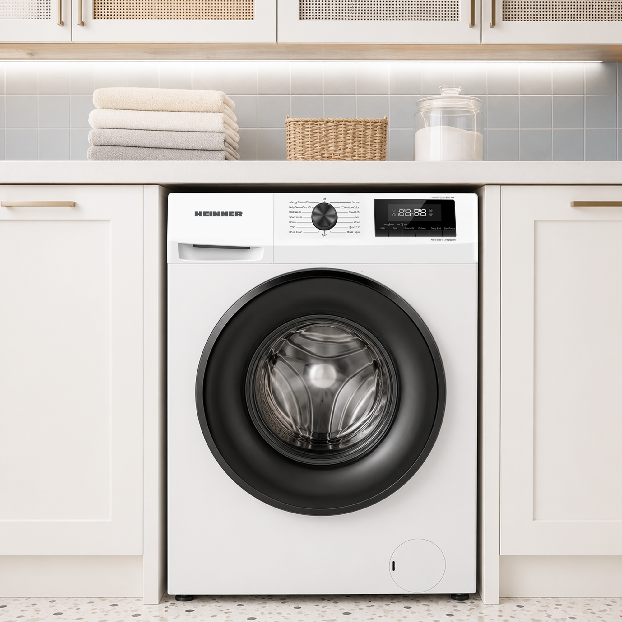 Un lave-linge blanc Heinner est placé sous un comptoir avec des serviettes pliées, un panier et un récipient en verre.