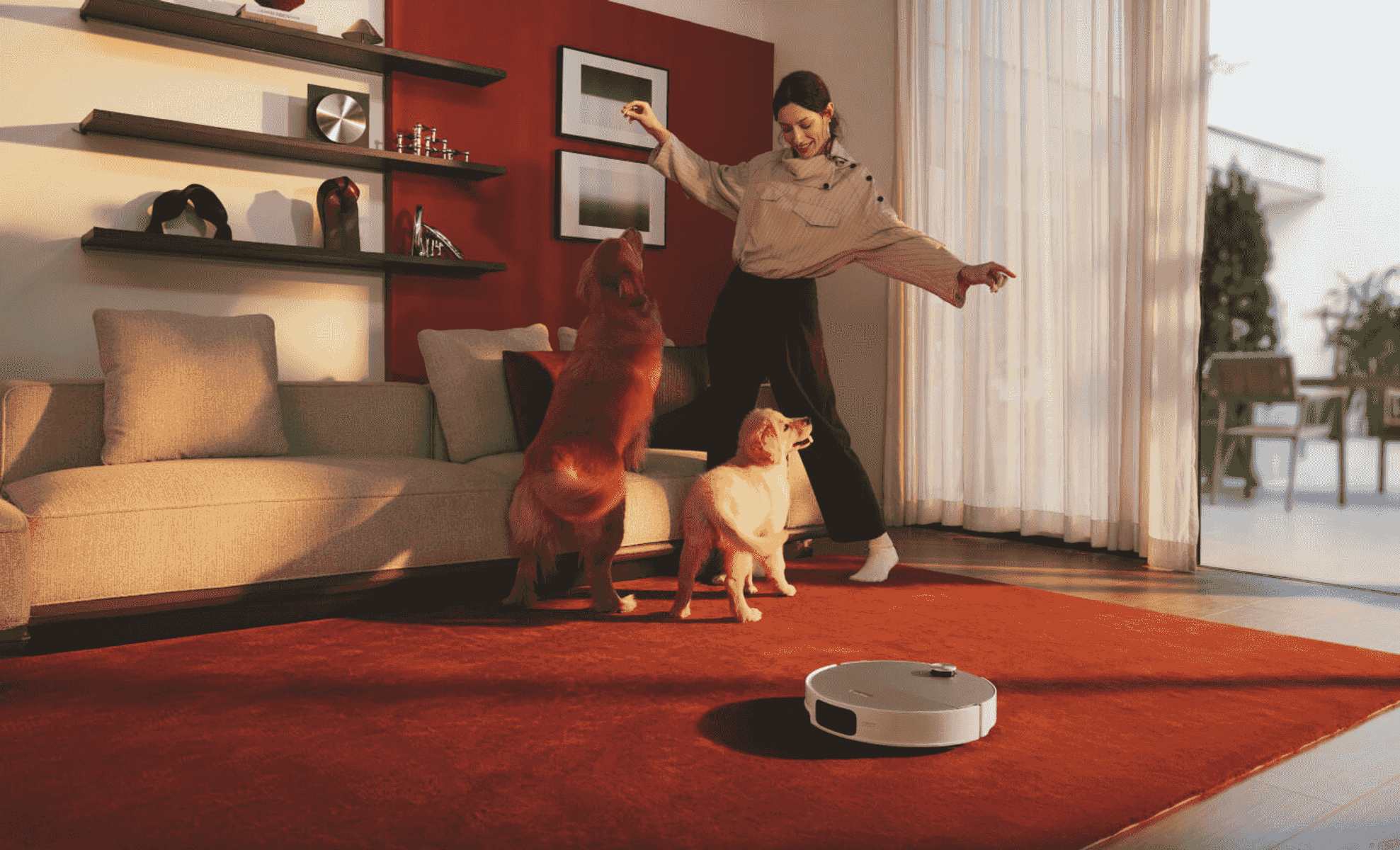 Una donna gioca con due cani su un tappeto rosso in salotto mentre un robot aspirapolvere si muove.