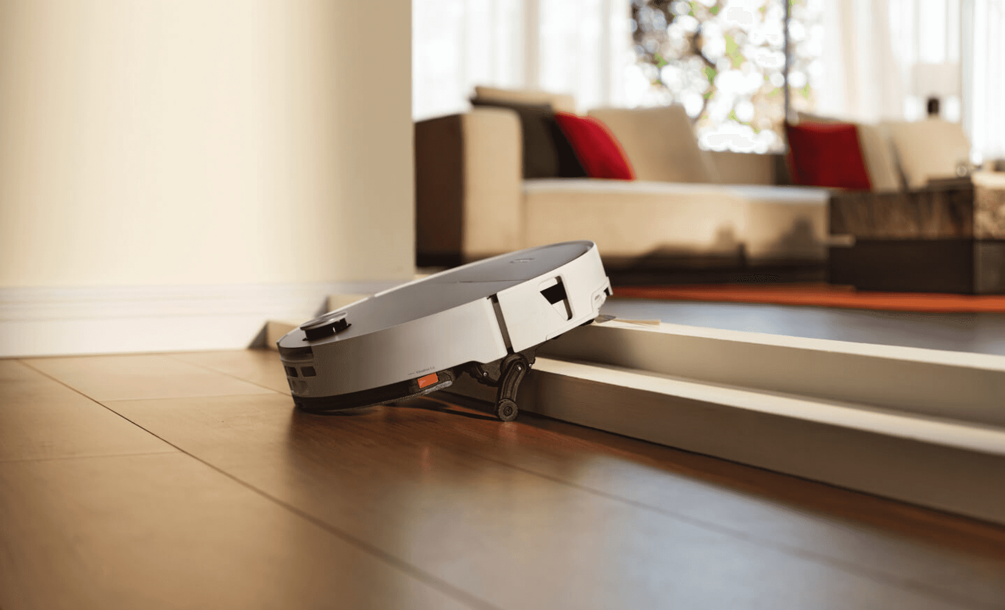 Un robot aspirapolvere bianco supera una soglia bassa su un pavimento in legno, con un soggiorno sfocato sullo sfondo.