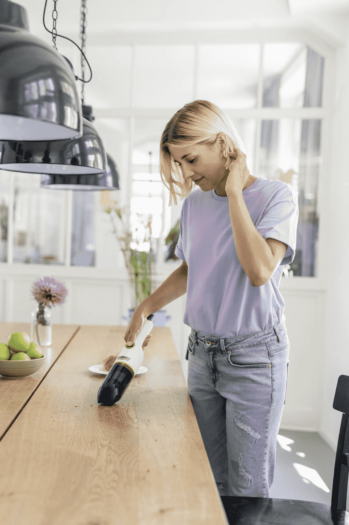 Donna bionda con maglietta viola e jeans aspira il tavolo della cucina con un aspirabriciole.