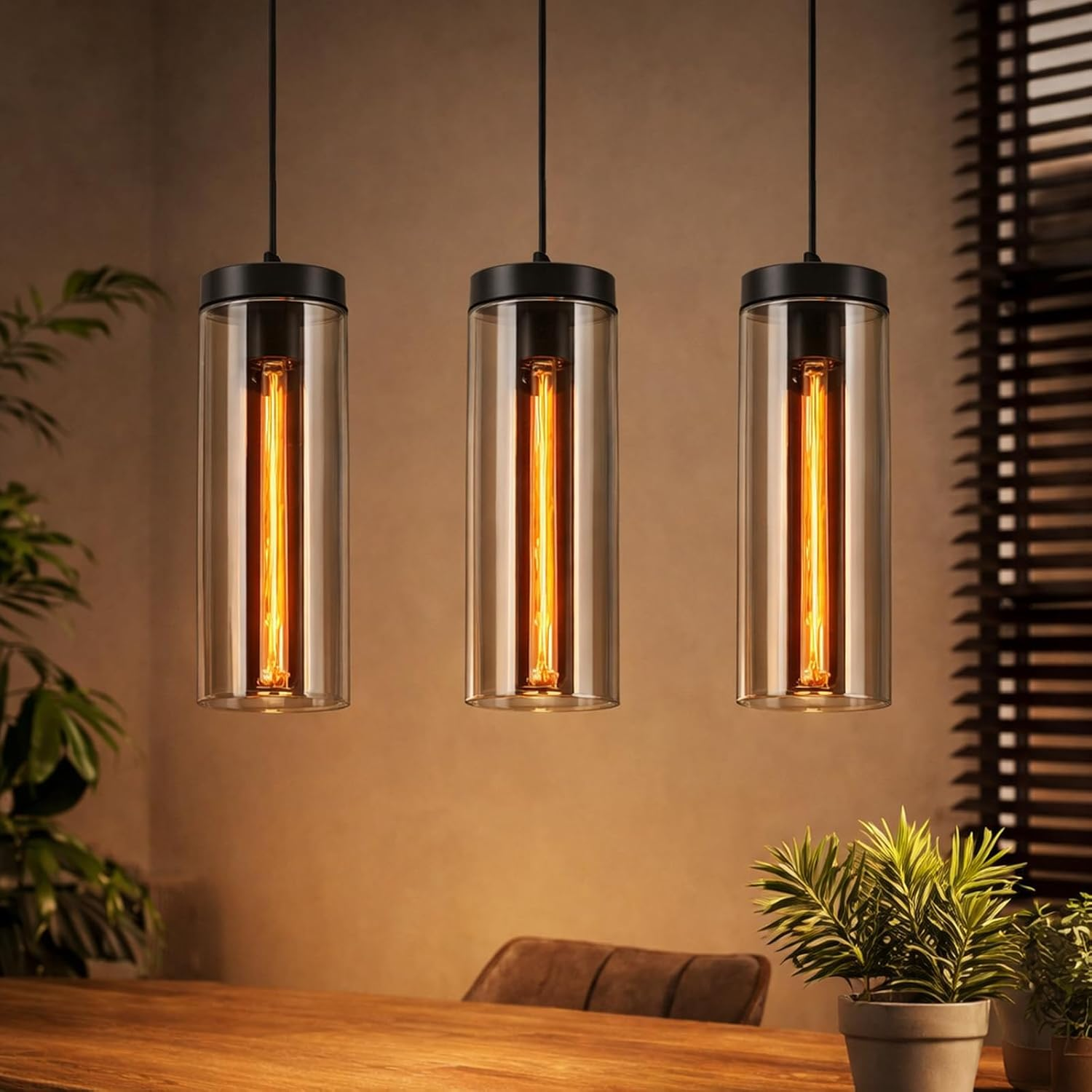 Drei Pendelleuchten mit zylindrischen Glasschirmen und glühenden Glühfadenlampen hängen über einem Holztisch.