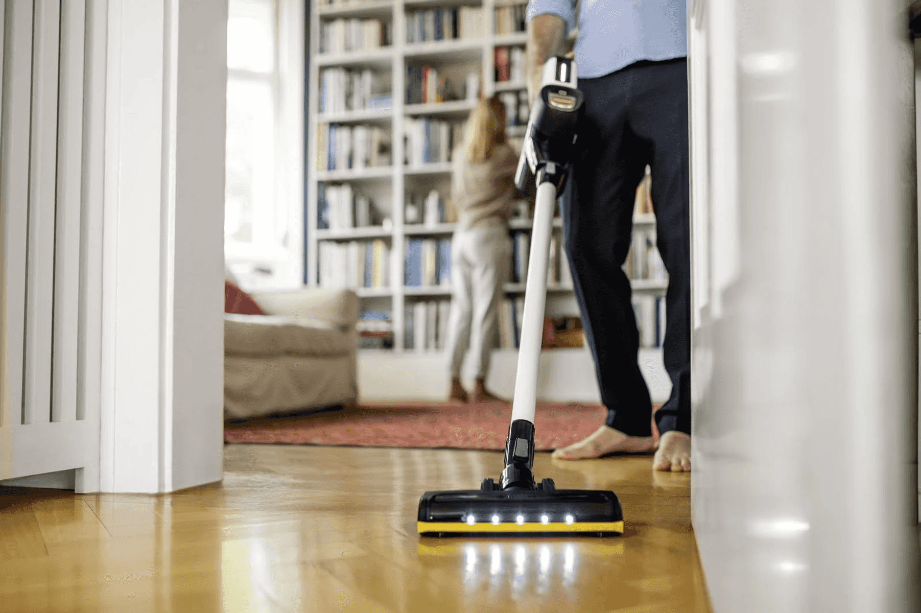 Un homme passe l'aspirateur sur un sol en bois avec un aspirateur sans fil dans un salon lumineux.