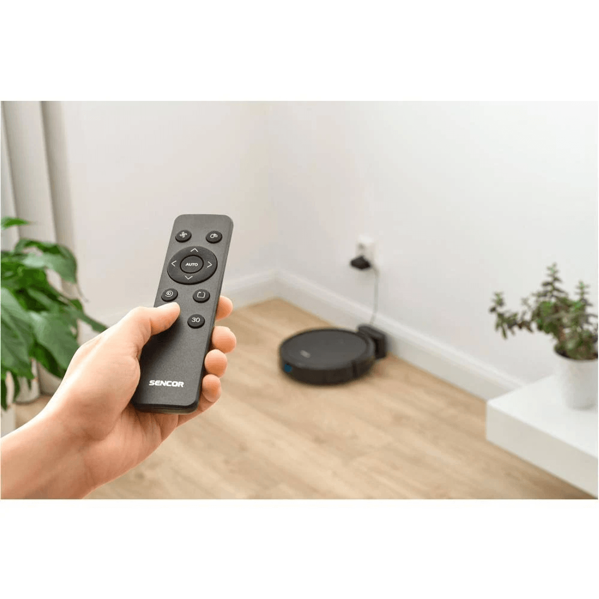 Una mano tiene un telecomando Sencor, controllando un aspirapolvere robot su un pavimento in legno.