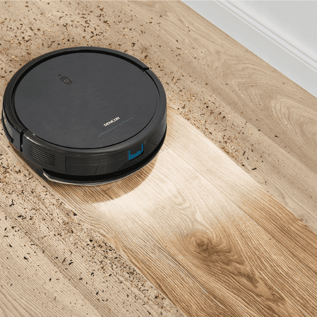 Un robot aspirapolvere Sencor nero pulisce un pavimento in legno parzialmente coperto di detriti.