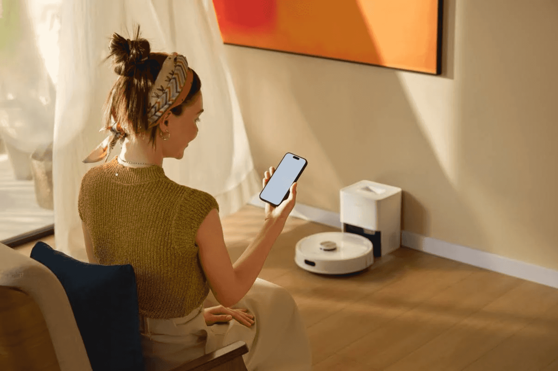 Una donna siede su una sedia, tiene uno smartphone e guarda un robot aspirapolvere in una stanza luminosa.