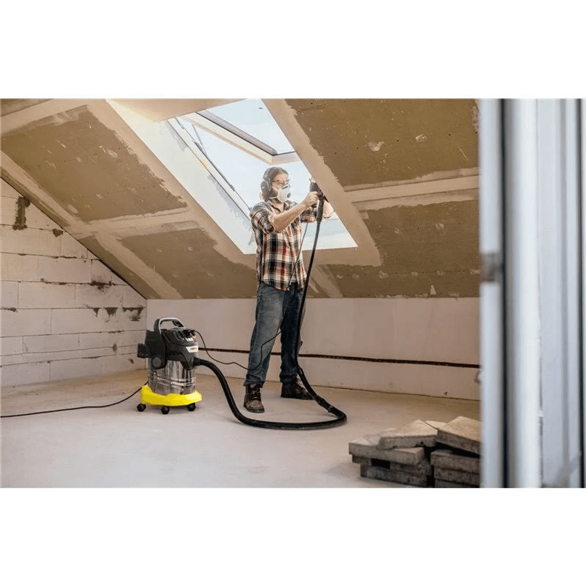 Un lavoratore con mascherina usa un elettroutensile e un aspirapolvere giallo in un cantiere in mansarda.