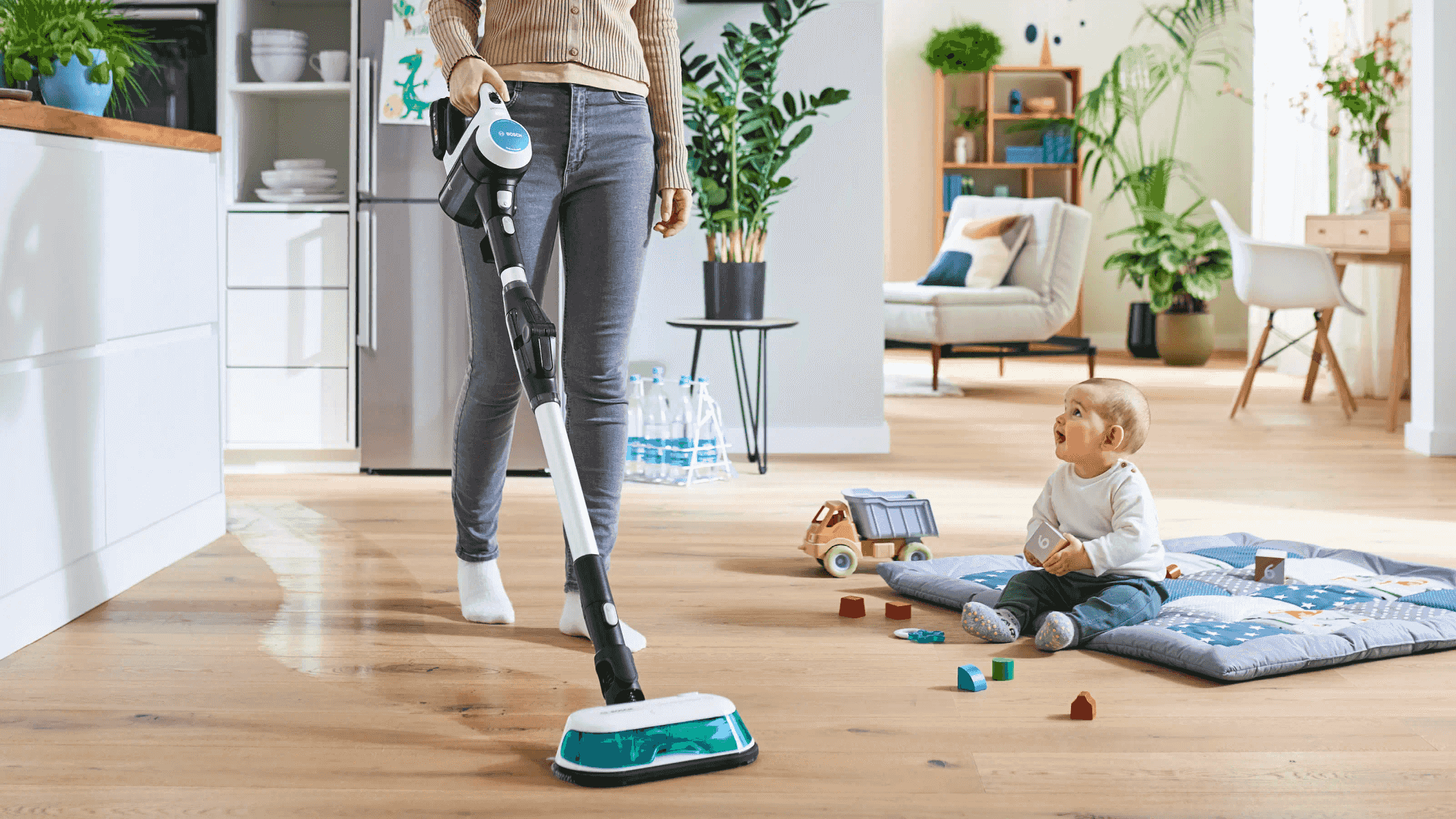 Una persona passa l'aspirapolvere senza fili sul pavimento in legno mentre un bambino gioca.