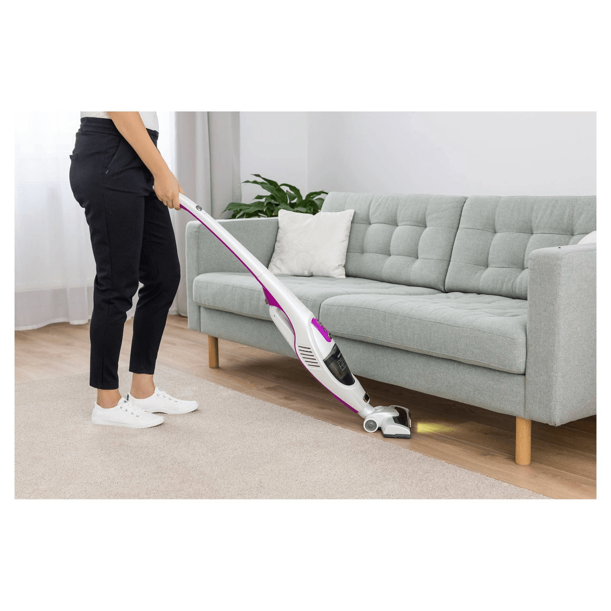 Eine Person in schwarzen Hosen benutzt einen weiß-lila Akkusauger neben einem grauen Sofa.