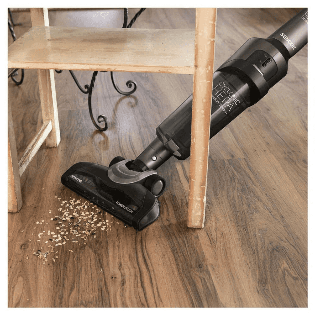 Un aspirapolvere nero pulisce i cereali sul pavimento di legno sotto un tavolo.