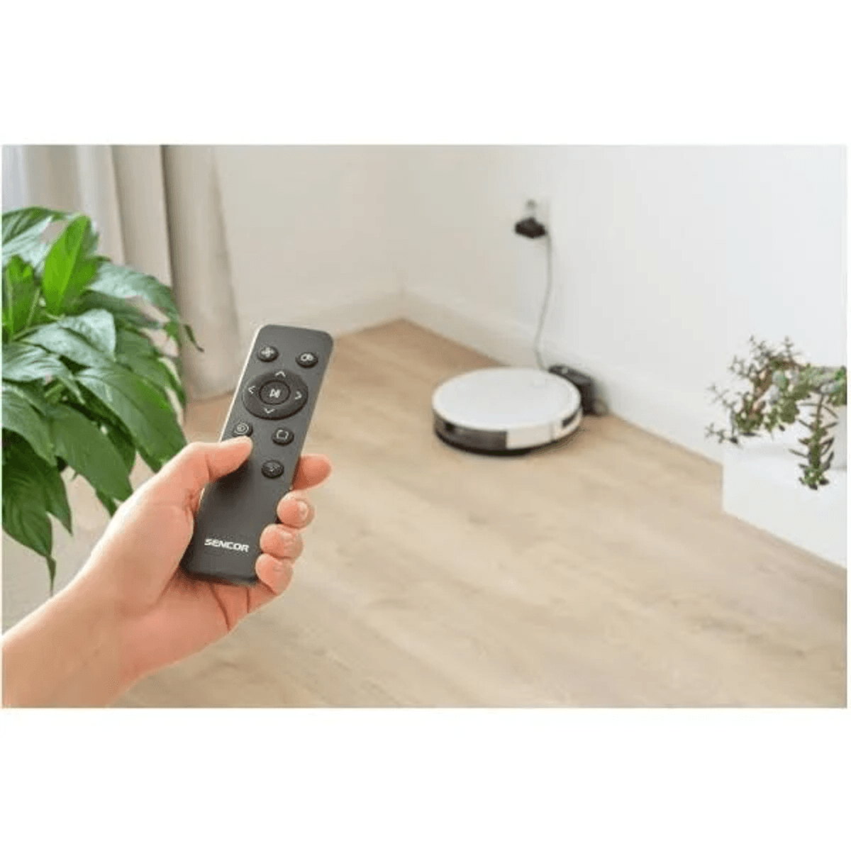 Una mano tiene un telecomando Sencor, mentre un robot aspirapolvere si trova sul pavimento in legno sullo sfondo.