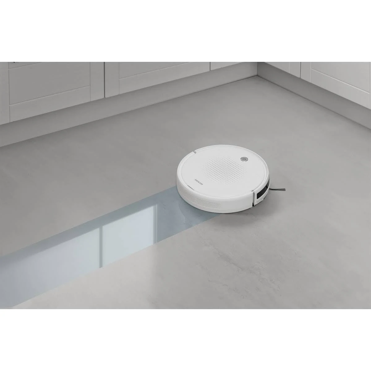 Un robot aspirapolvere bianco pulisce un pavimento in cemento in una cucina moderna con mobili grigi.