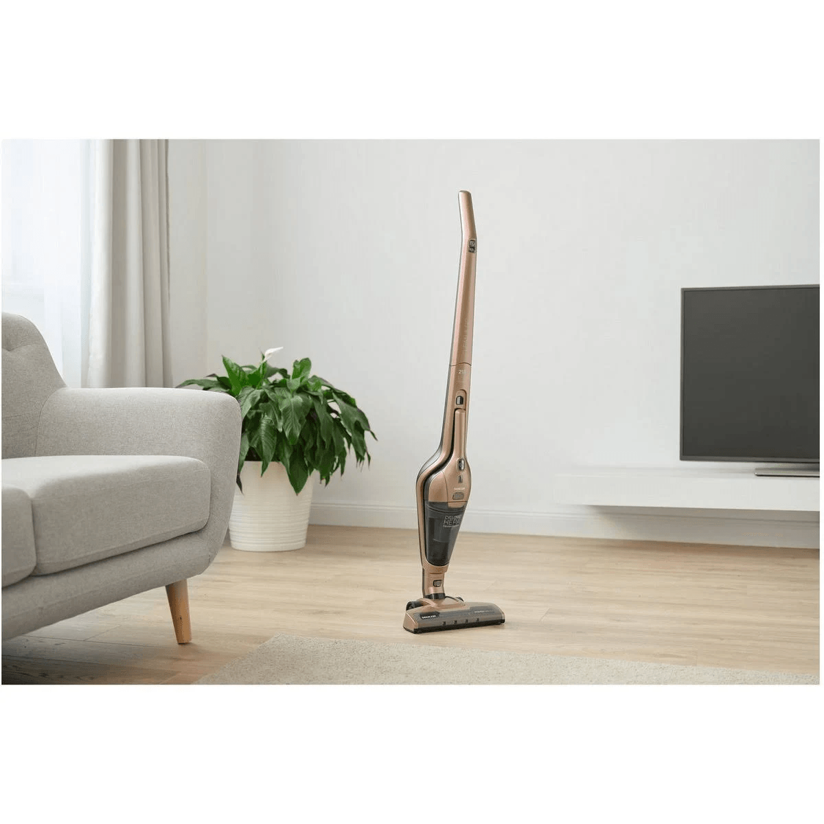 Un aspirapolvere senza fili bronzo si erge su un pavimento in legno in un soggiorno con divano e pianta.