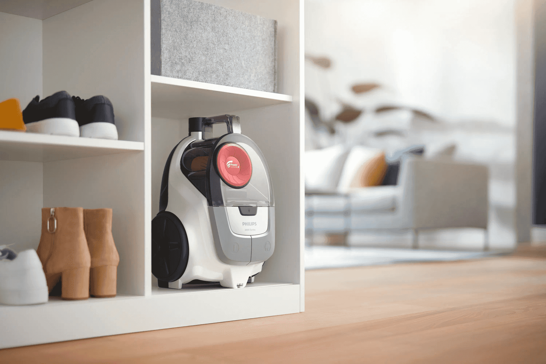 Ein weiß-grauer Philips Staubsauger steht in einem Regal neben Schuhen, im Hintergrund ein Wohnzimmer.
