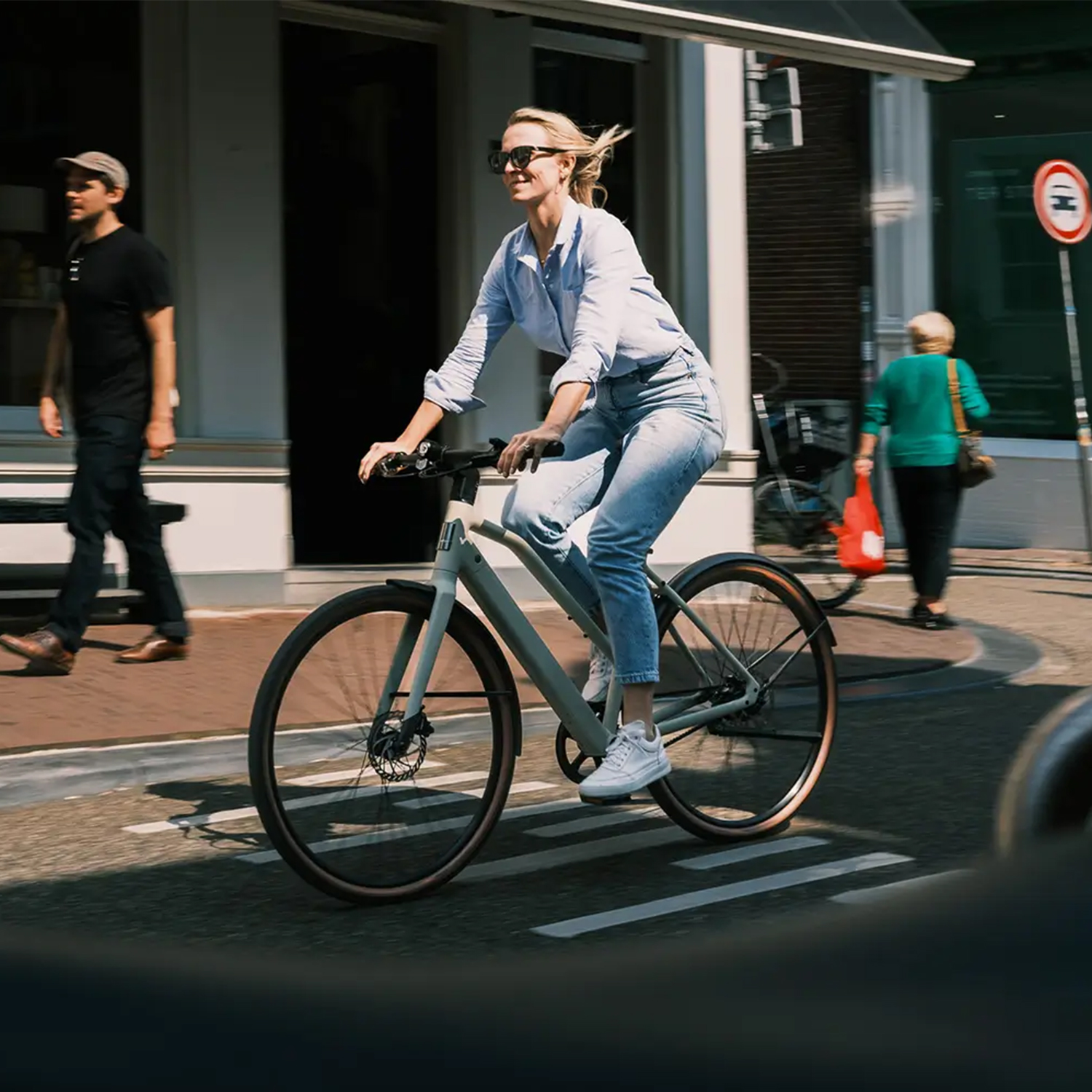 Eine blonde Frau in Jeans und hellem Hemd fährt ein helles Fahrrad auf einer Straße, mit Passanten im Hintergrund.