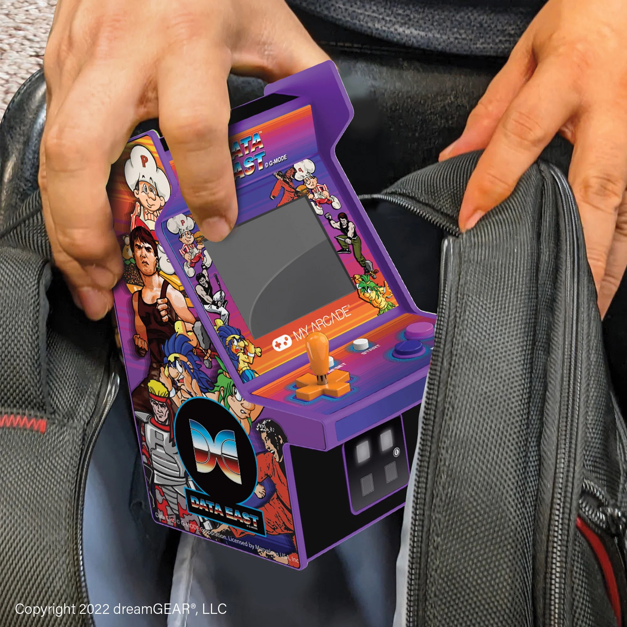 Eine Person steckt einen violetten Retro-Mini-Spielautomaten mit Spielefiguren in einen Rucksack.