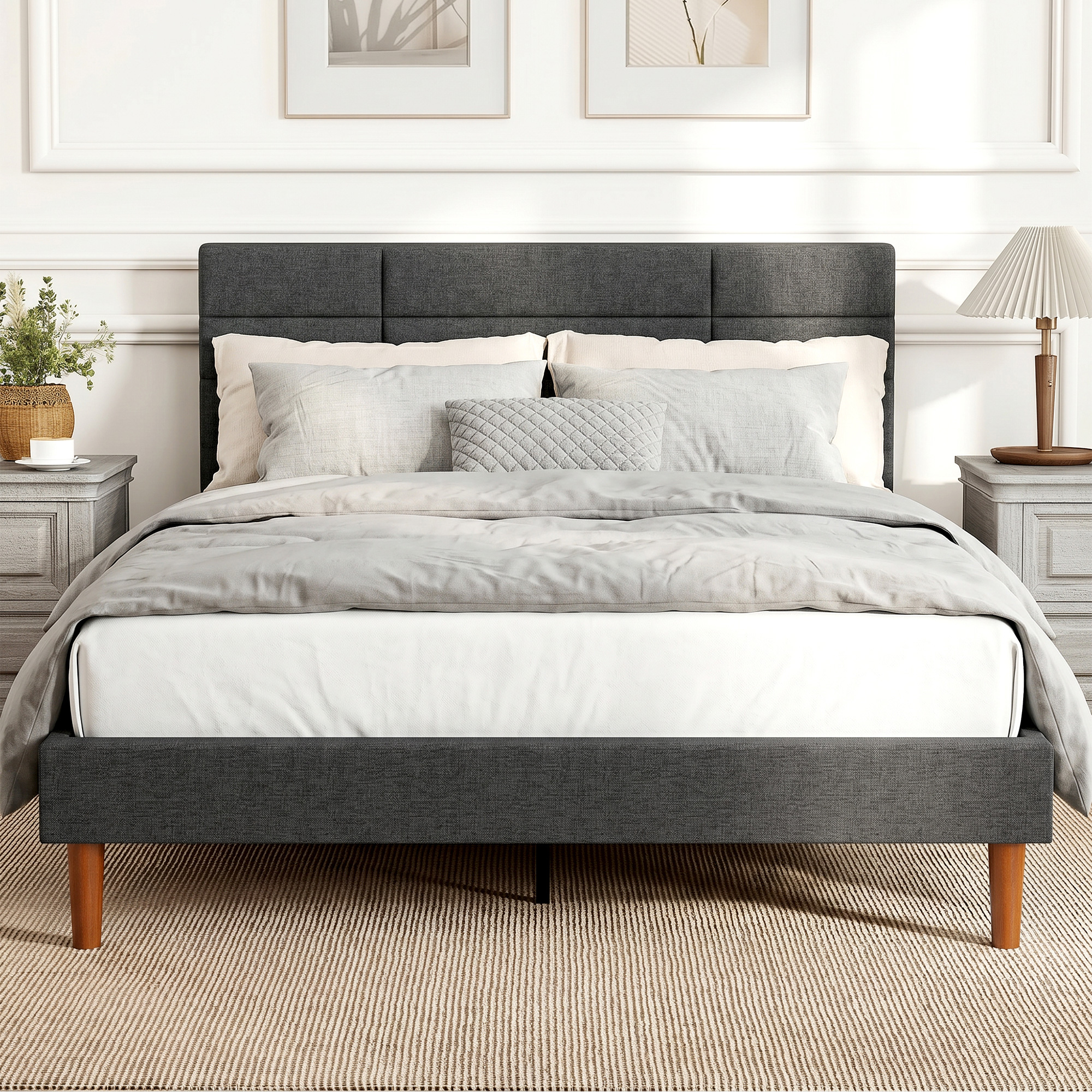 Una cama gris con patas de madera y ropa de cama gris, flanqueada por mesitas de noche con una planta y una lámpara.