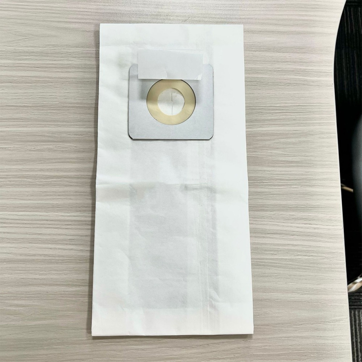 Ein weißer Staubsaugerbeutel mit einer quadratischen Kartonhalterung und einer zentralen Öffnung auf Holztisch.
