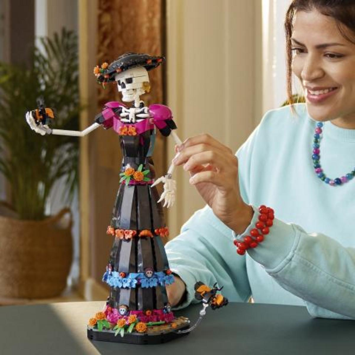 Açık mavi kazak giymiş gülümseyen bir kadın, renkli elbiseli bir LEGO iskelet figürü tutuyor.