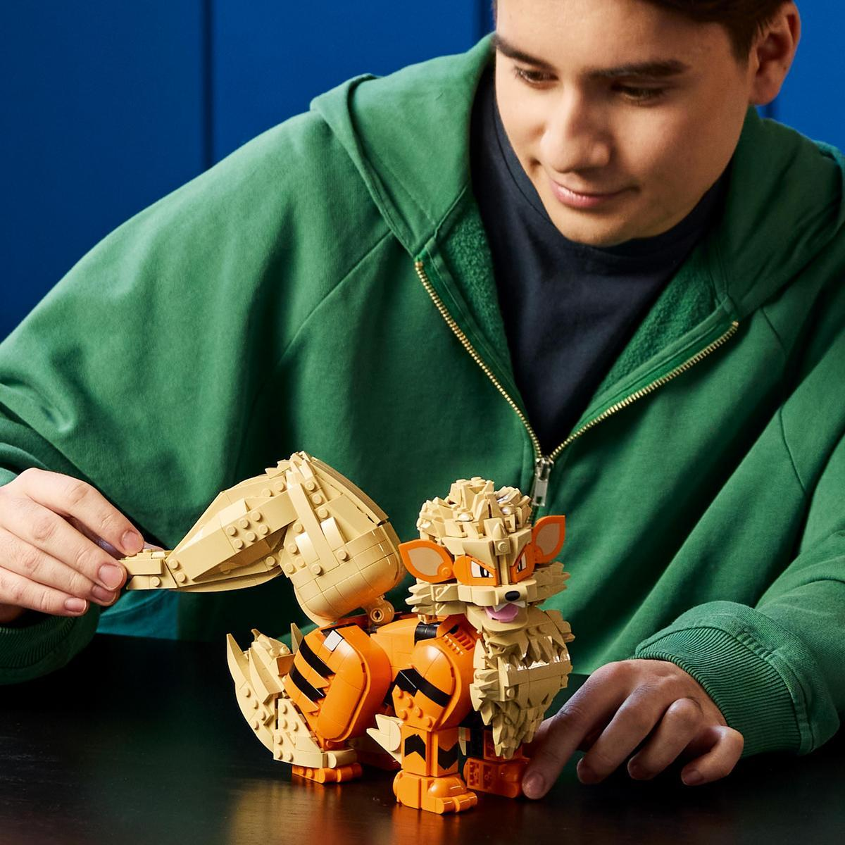Yeşil kapüşonlu genç bir adam masada turuncu ve bej renkli bir Lego hayvan modeli yapıyor.