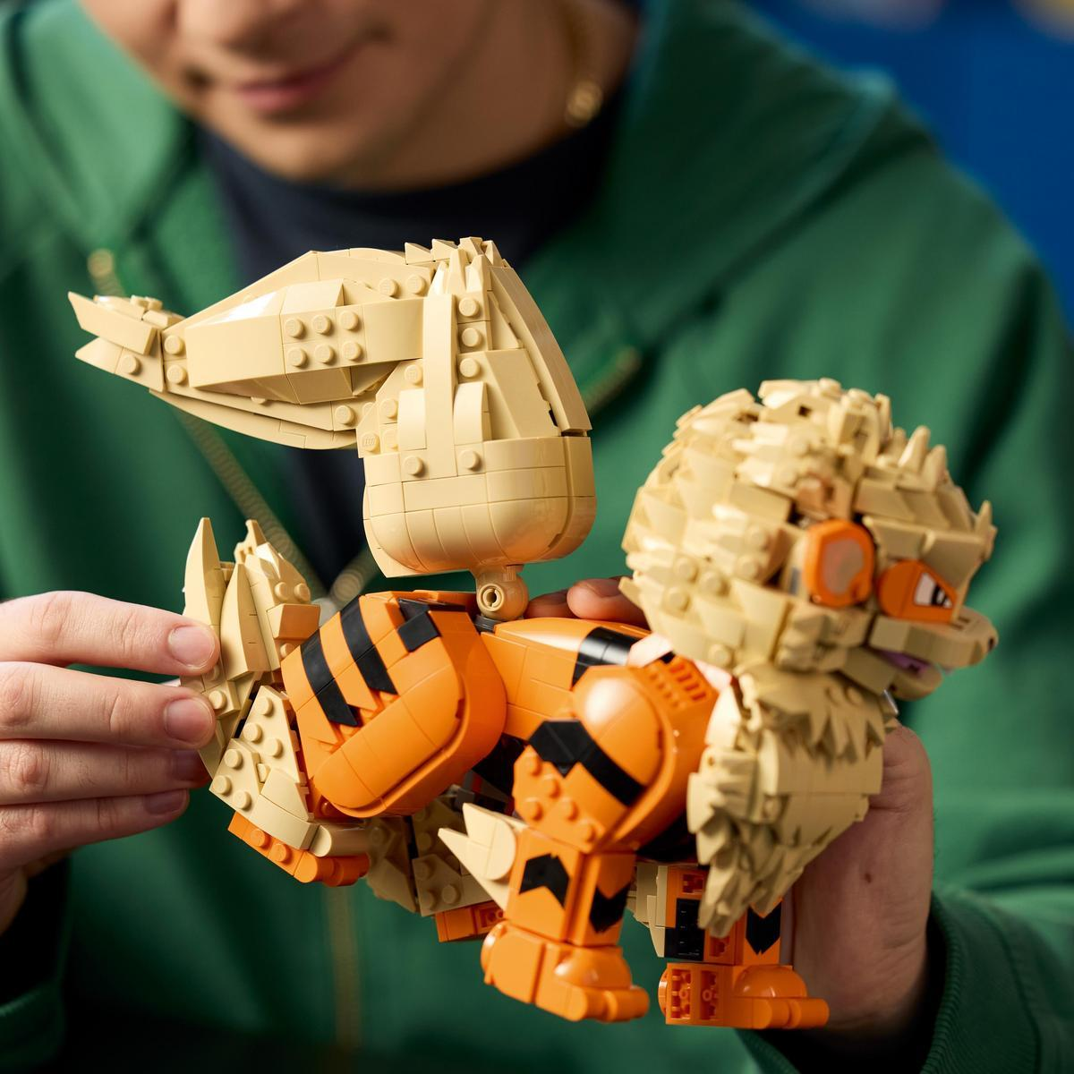 Bir kişi büyük tüylü bacakları ve büyük kuyruğu olan turuncu ve siyah bir Lego hayvan figürü inşa ediyor.