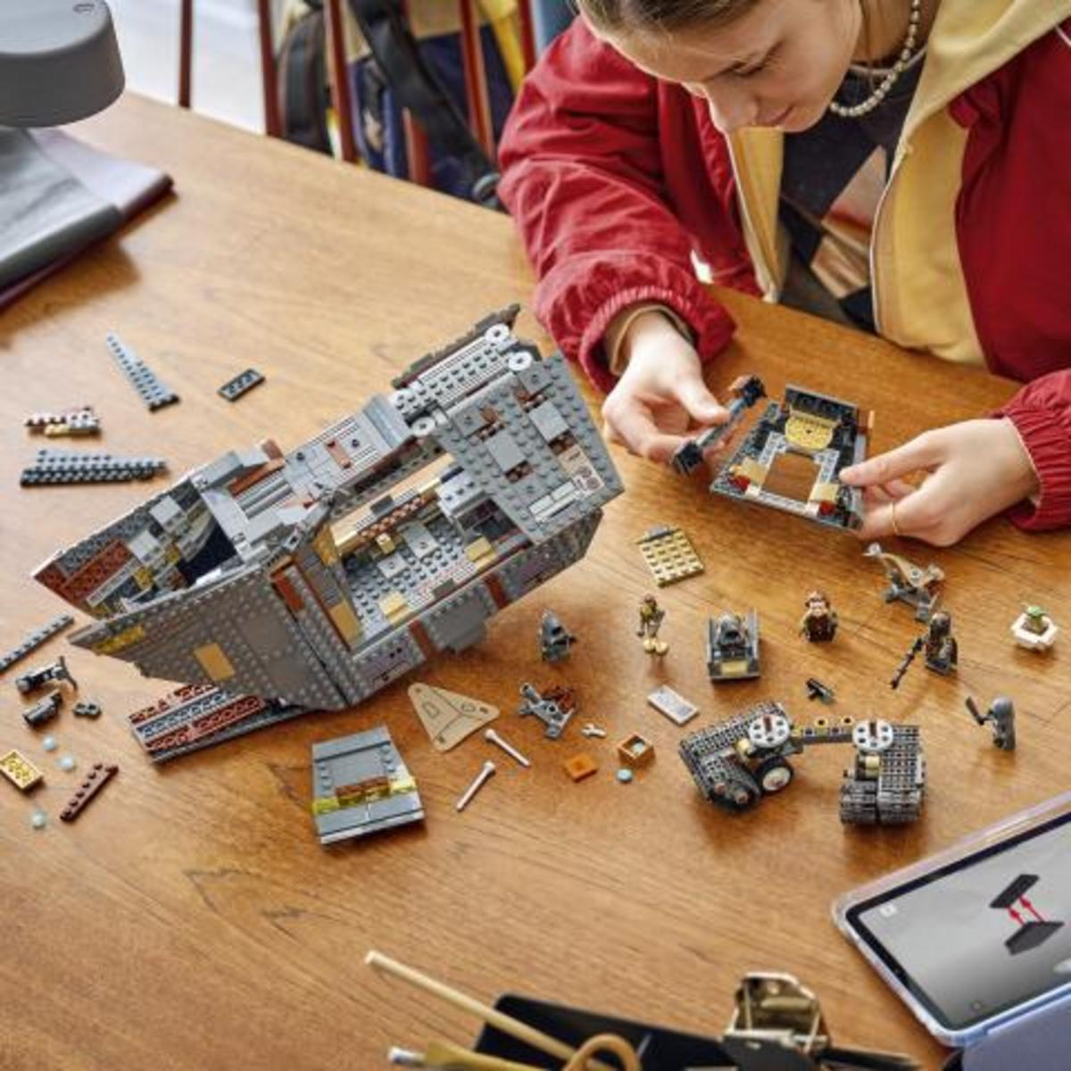 Una persona construye un gran modelo de nave espacial Lego en una mesa de madera, rodeada de piezas y minifiguras.