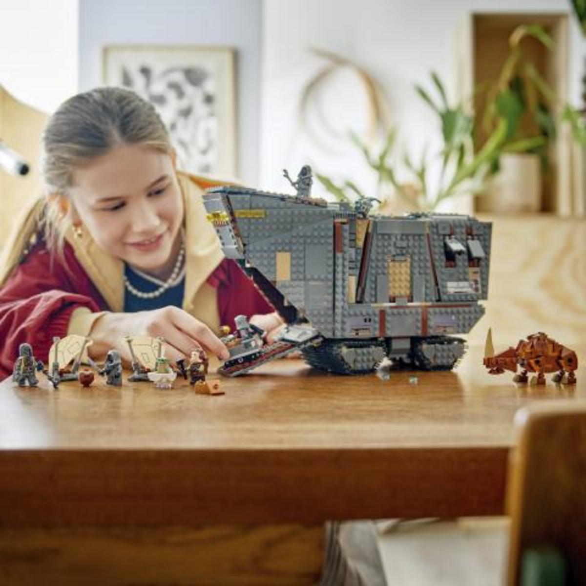 Una niña construye un gran modelo de nave LEGO con minifiguras y una criatura en una mesa de madera.