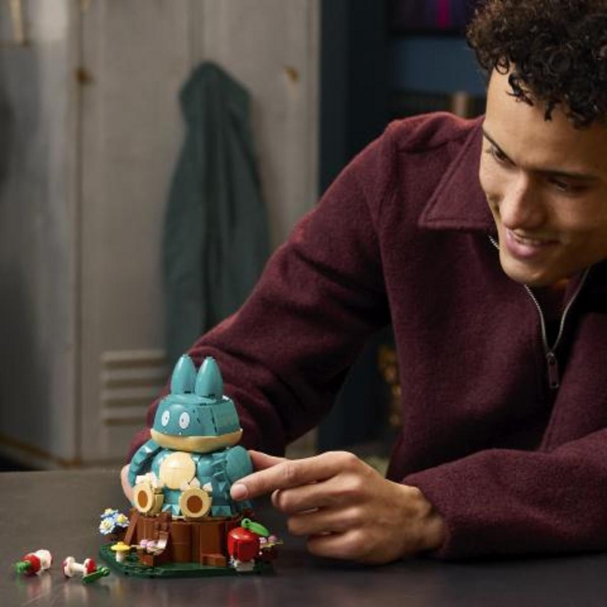 Un joven sonriente con una chaqueta granate construyendo un personaje azul de Lego en un tronco.