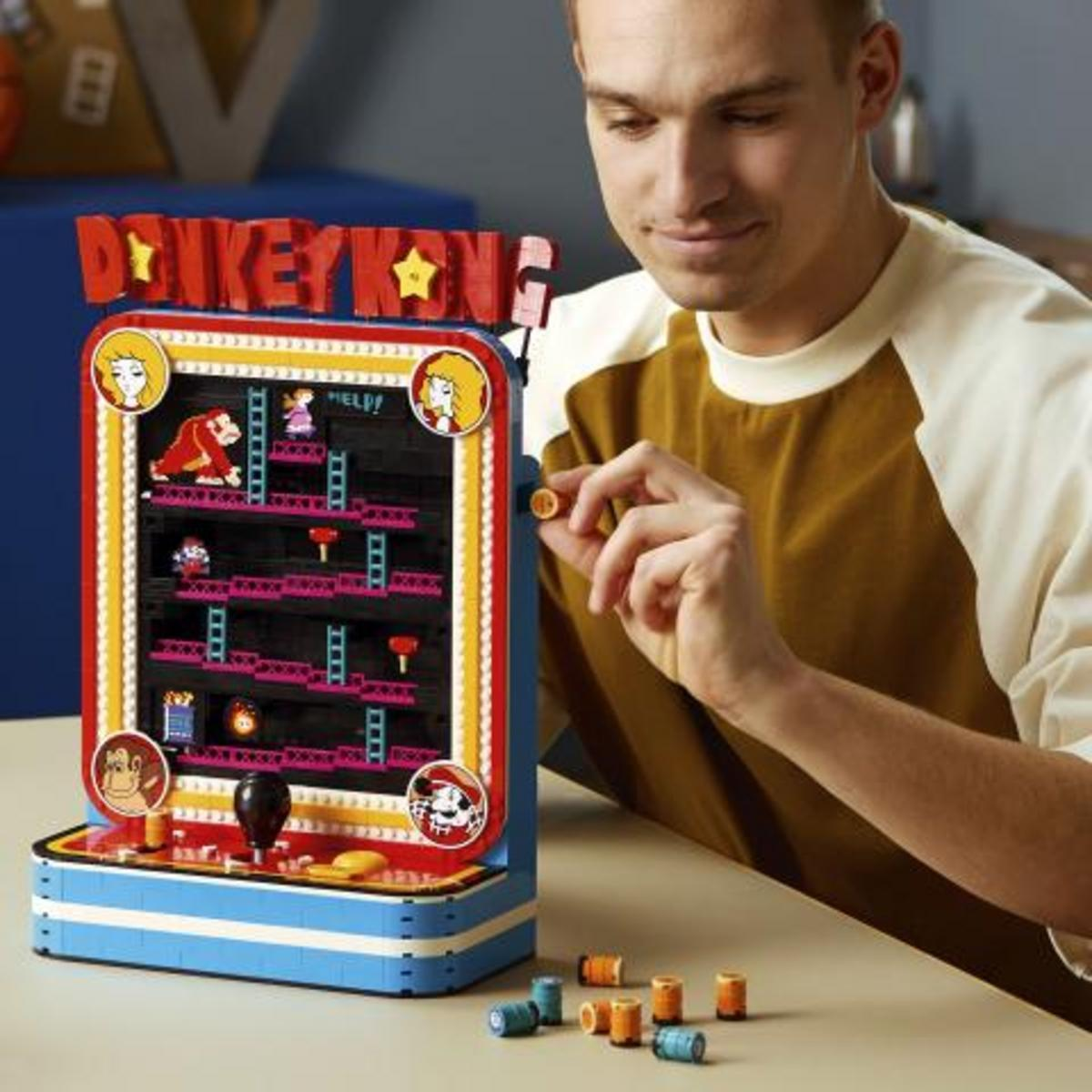 Een man bouwt een kleurrijk Donkey Kong arcadespel van LEGO-blokjes op een tafel.