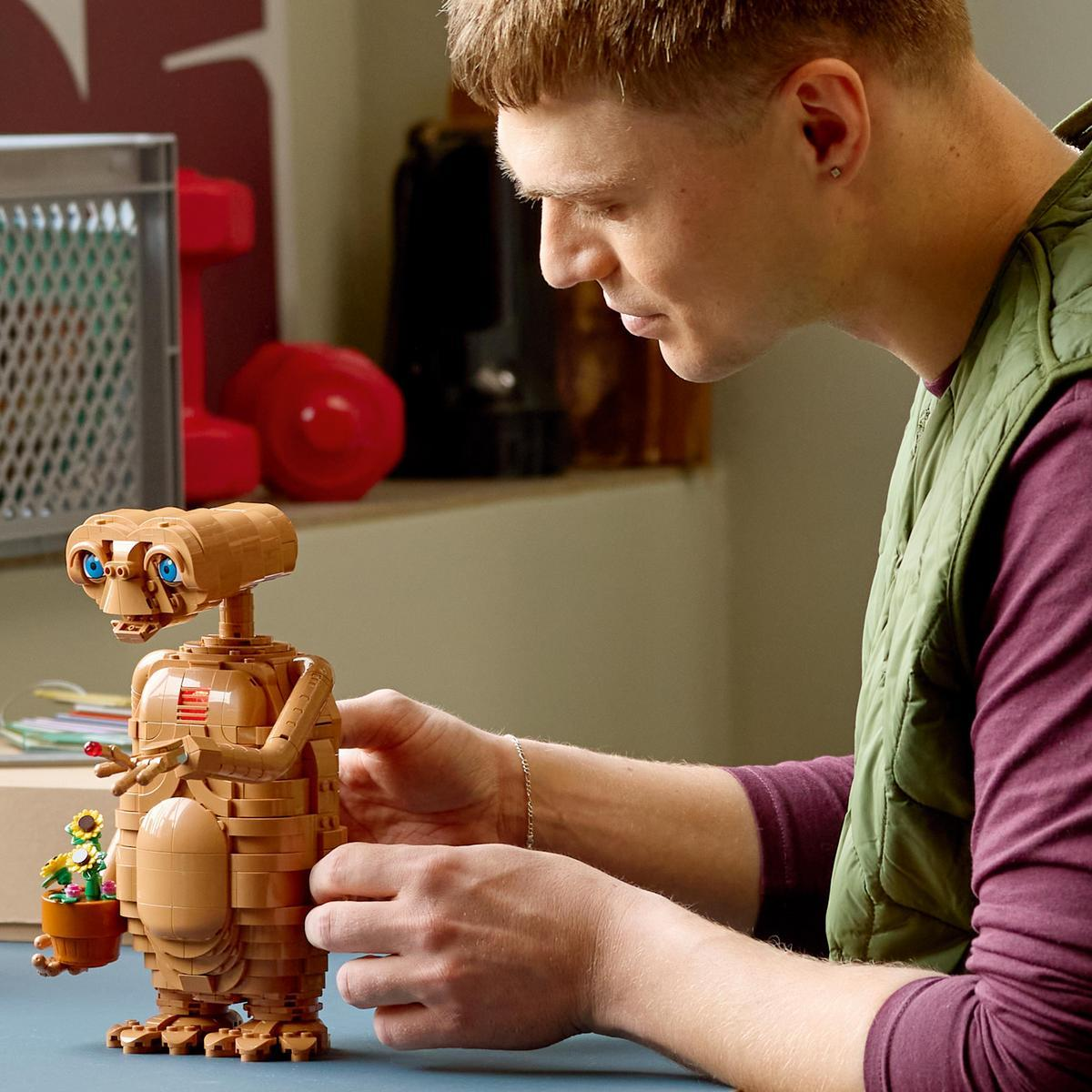 Un hombre construye una figura LEGO E.T. marrón que sostiene una maceta con flores en una mesa.