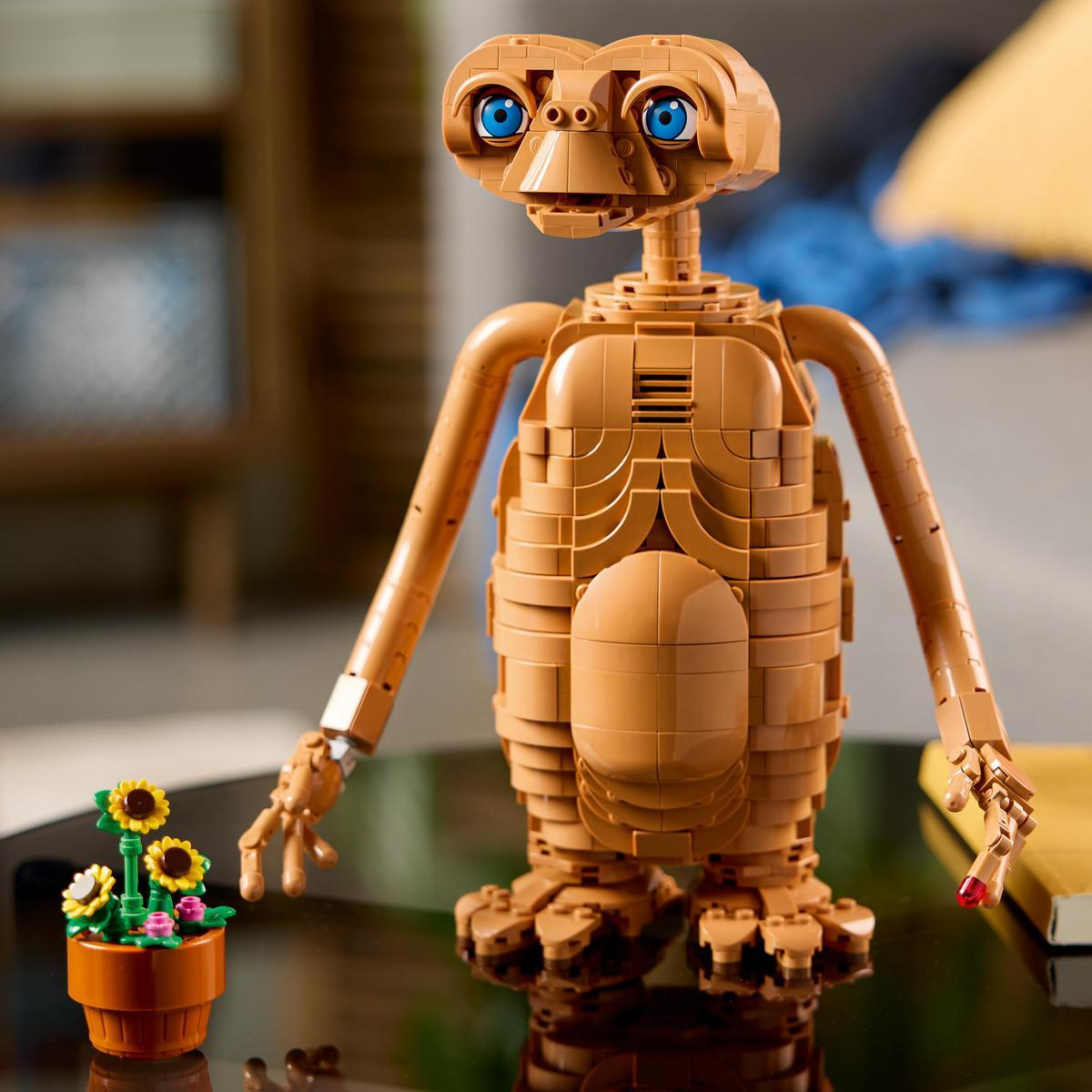 Una figura de E.T. hecha de Lego sobre una mesa de vidrio junto a una pequeña planta con flores amarillas.