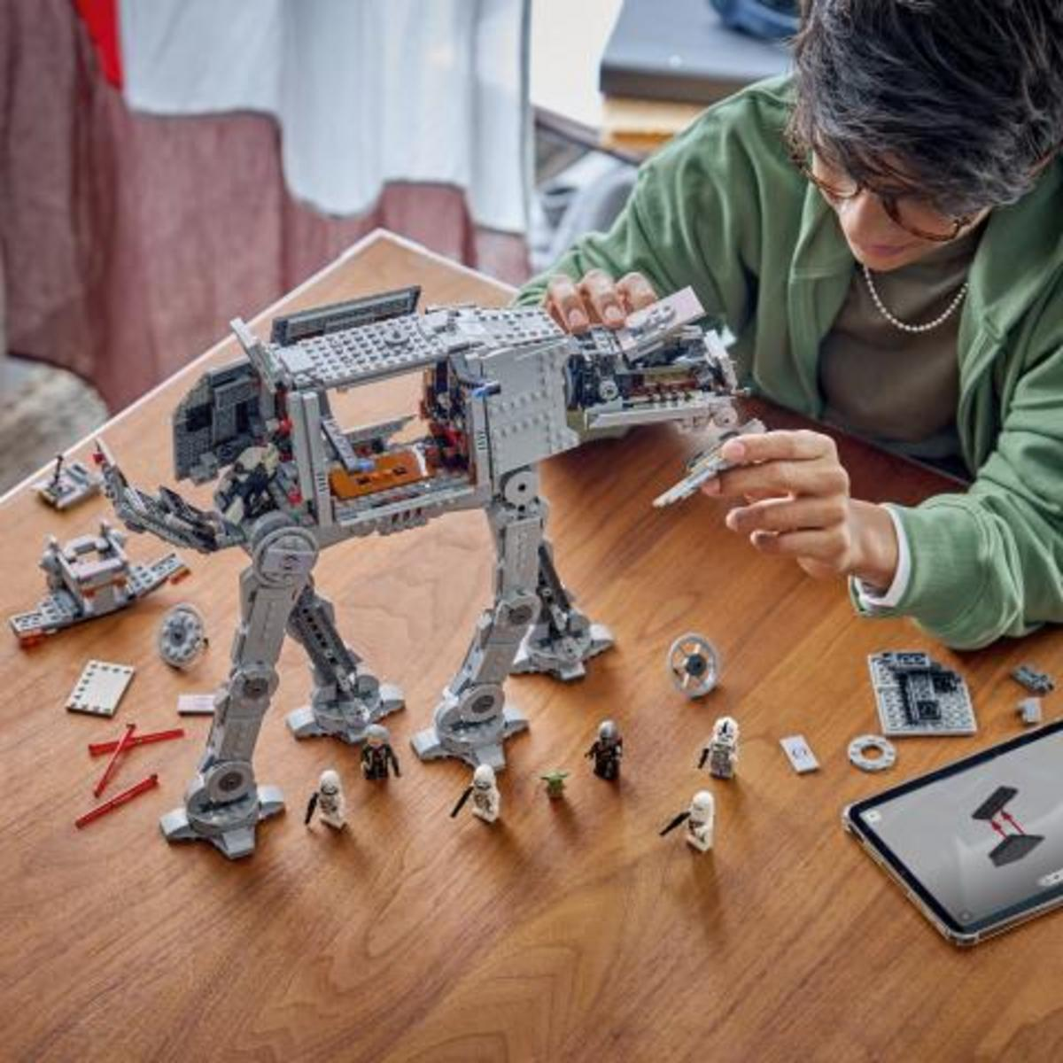 Una persona con sudadera verde monta un gran caminante LEGO AT-ST en una mesa de madera, rodeada de minifiguras y una tableta.