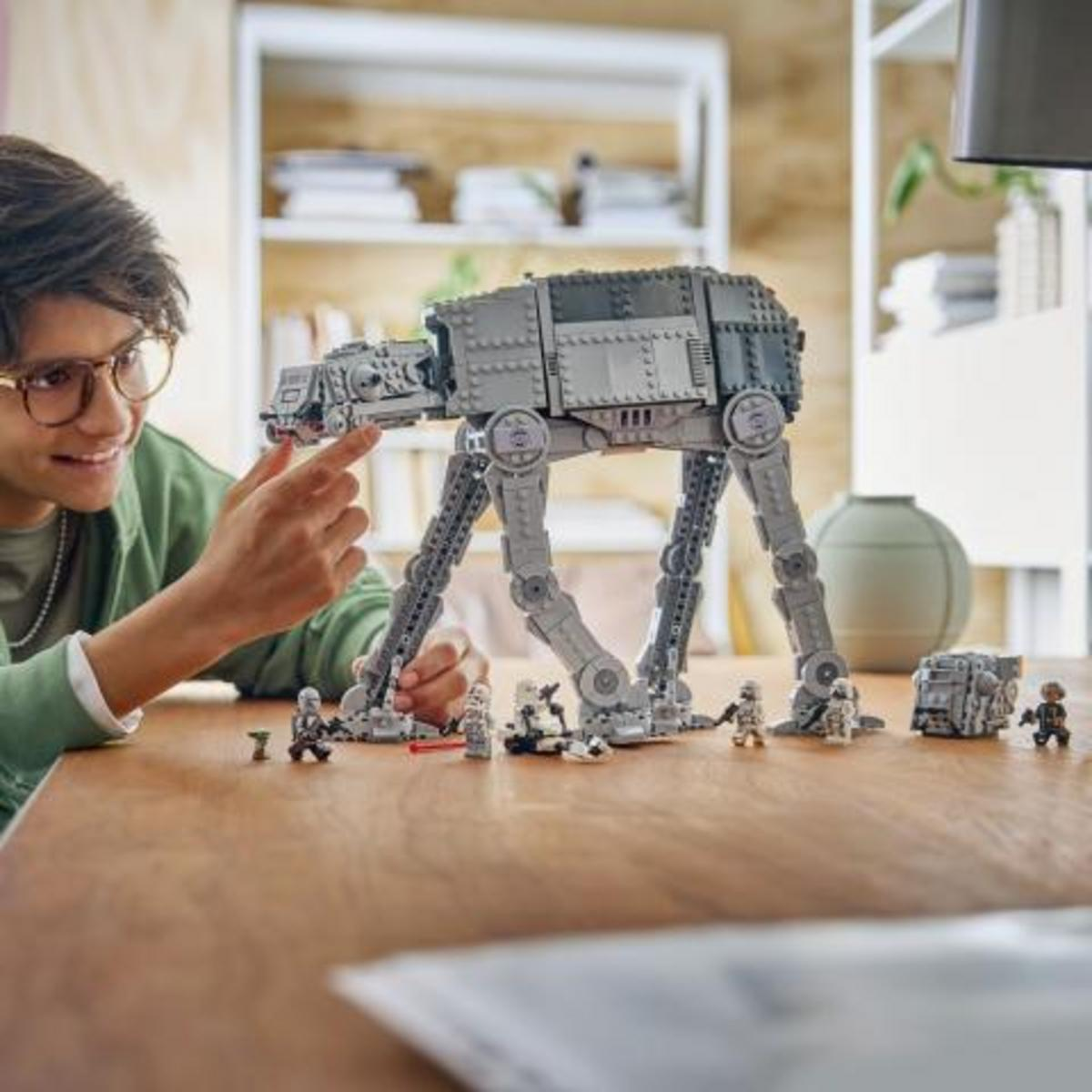 Un niño con gafas construye un gran modelo Lego AT-ST gris en una mesa de madera, rodeado de pequeñas figuras.