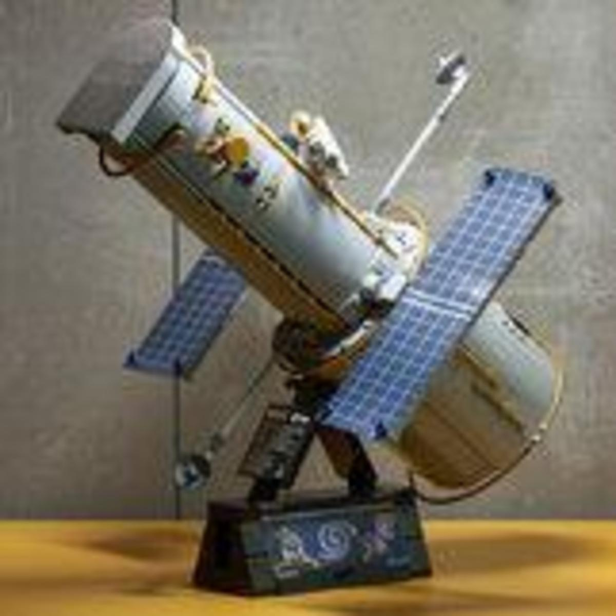 Un modelo detallado del Telescopio Espacial Hubble con paneles solares azules en un soporte ante una pared gris.