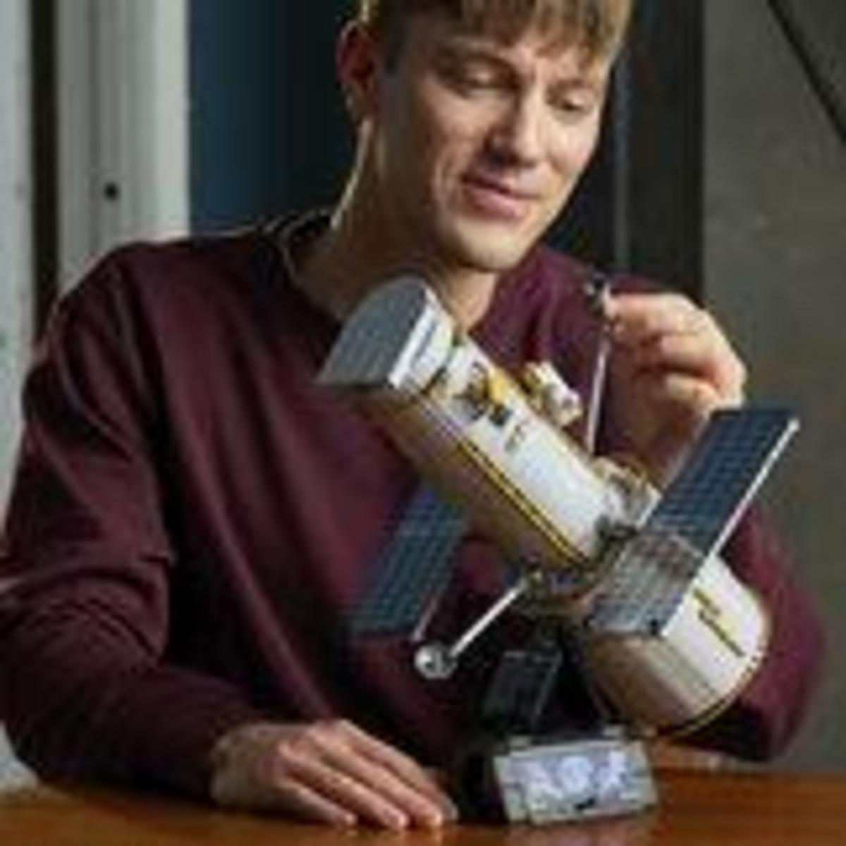 Un joven sonriente con suéter granate construye cuidadosamente un modelo detallado del Telescopio Espacial Hubble.