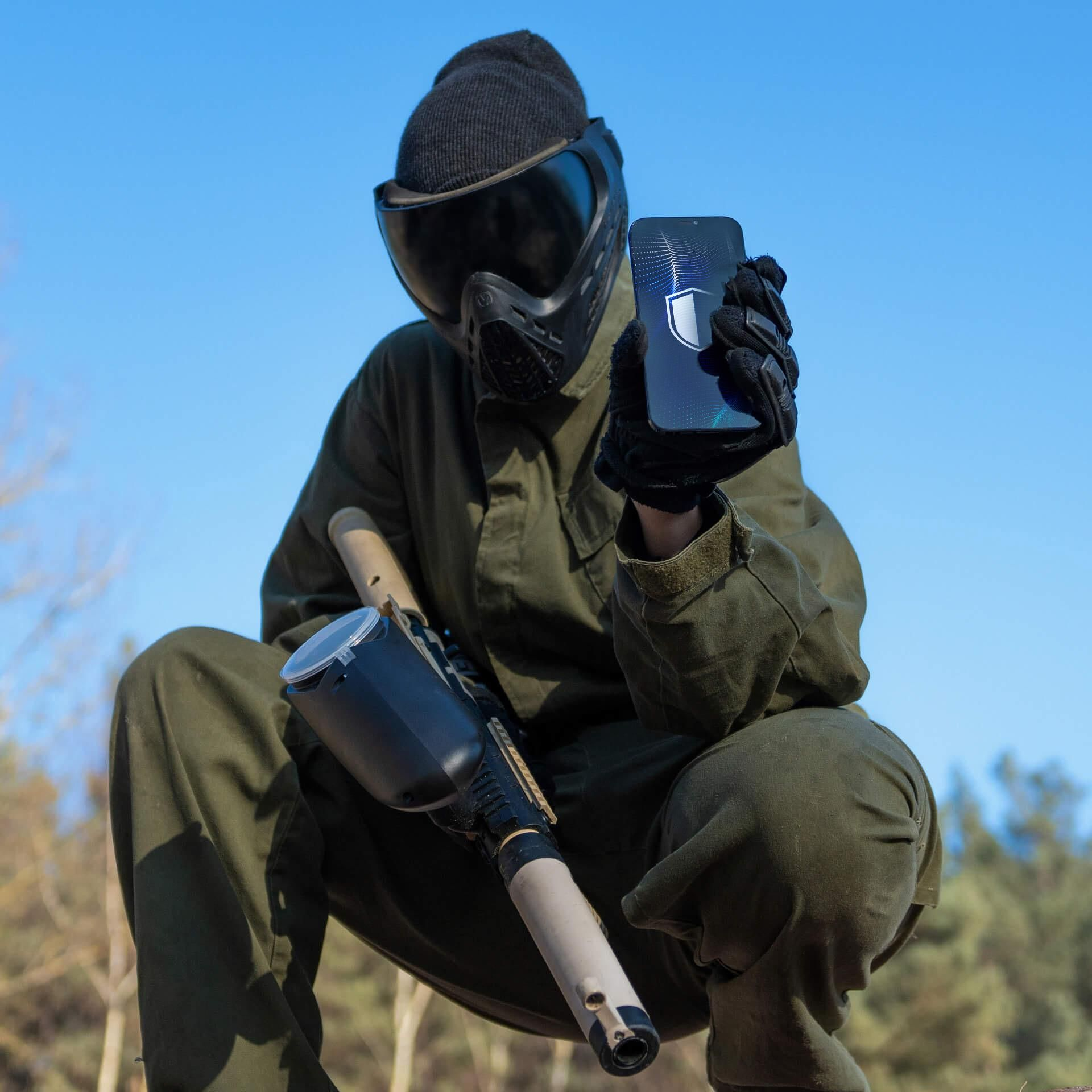 Una persona in verde con maschera protettiva e berretto è accovacciata all'aperto e mostra uno smartphone.